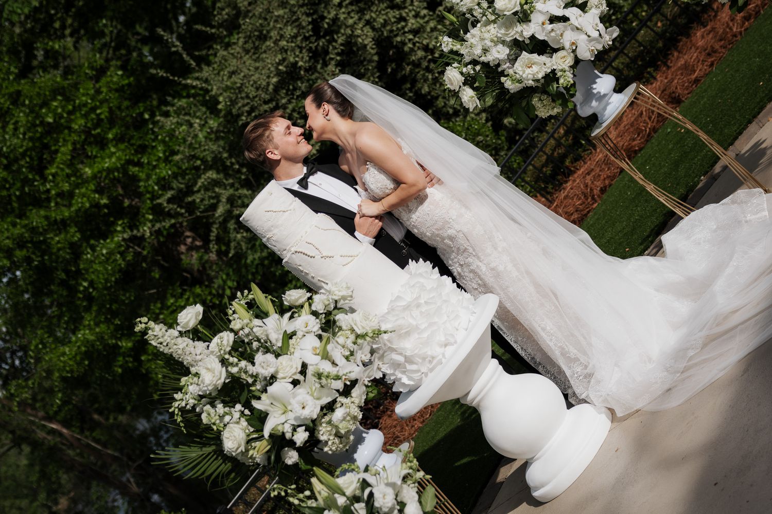 Bride and groom share a kiss at outdoor wedding altar surrounded by white floral arrangements.