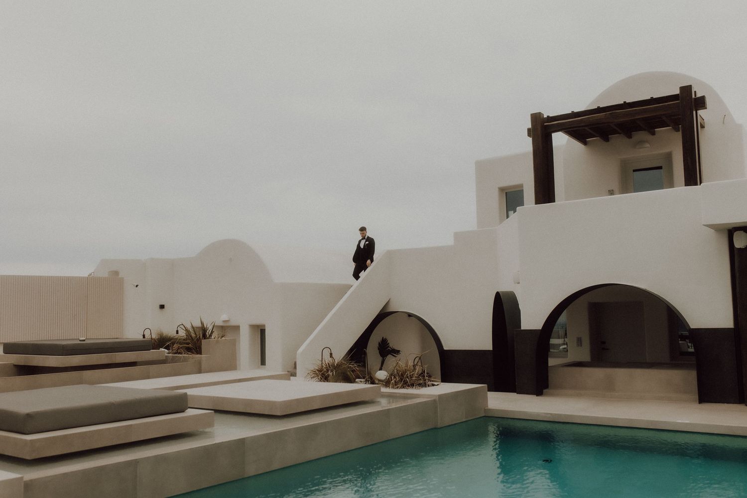 A minimalist white architectural space features a turquoise pool under an overcast sky.