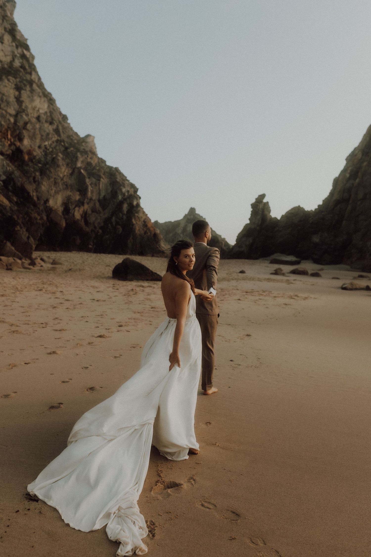 A couple in wedding attire walks along a sandy beach with dramatic rock formations.