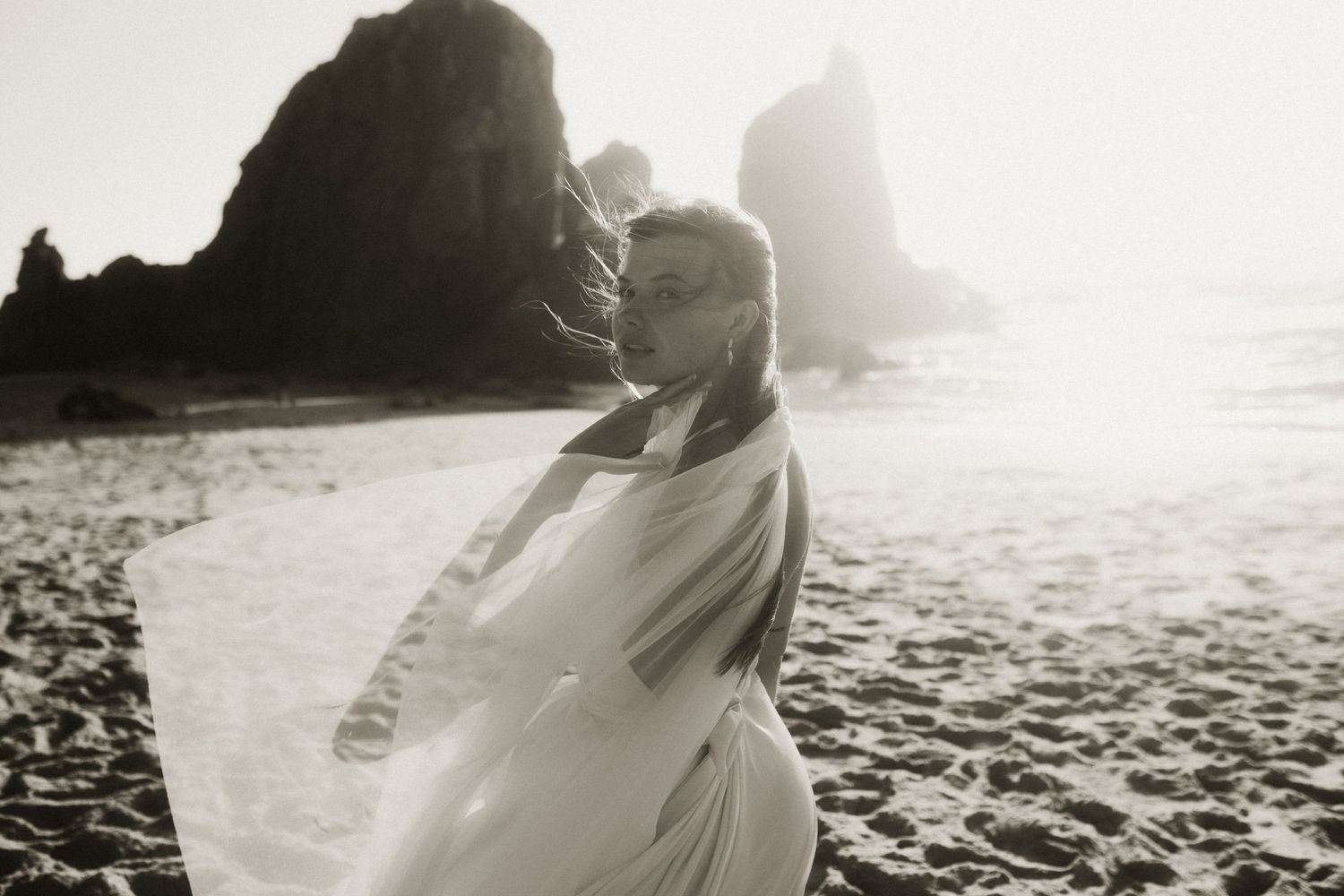 Sunlit wedding couple embraces on a sandy beach with waves crashing in the background.