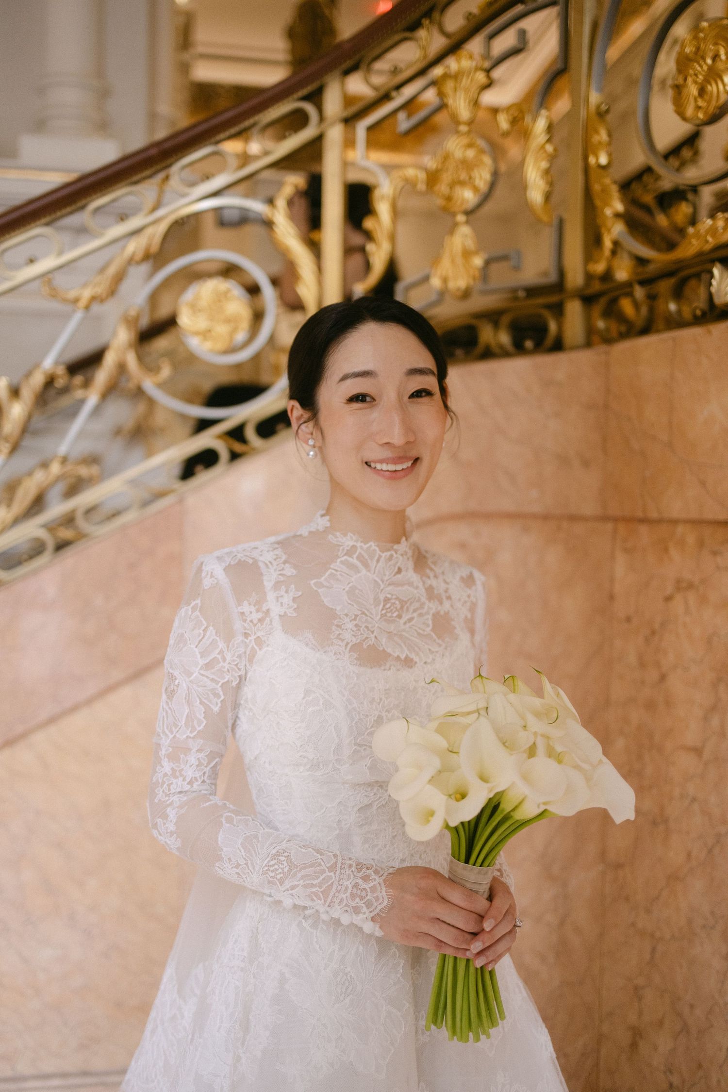 A bride in an elegant white lace wedding dress holds a white floral bouquet while standing in an ornate venue.