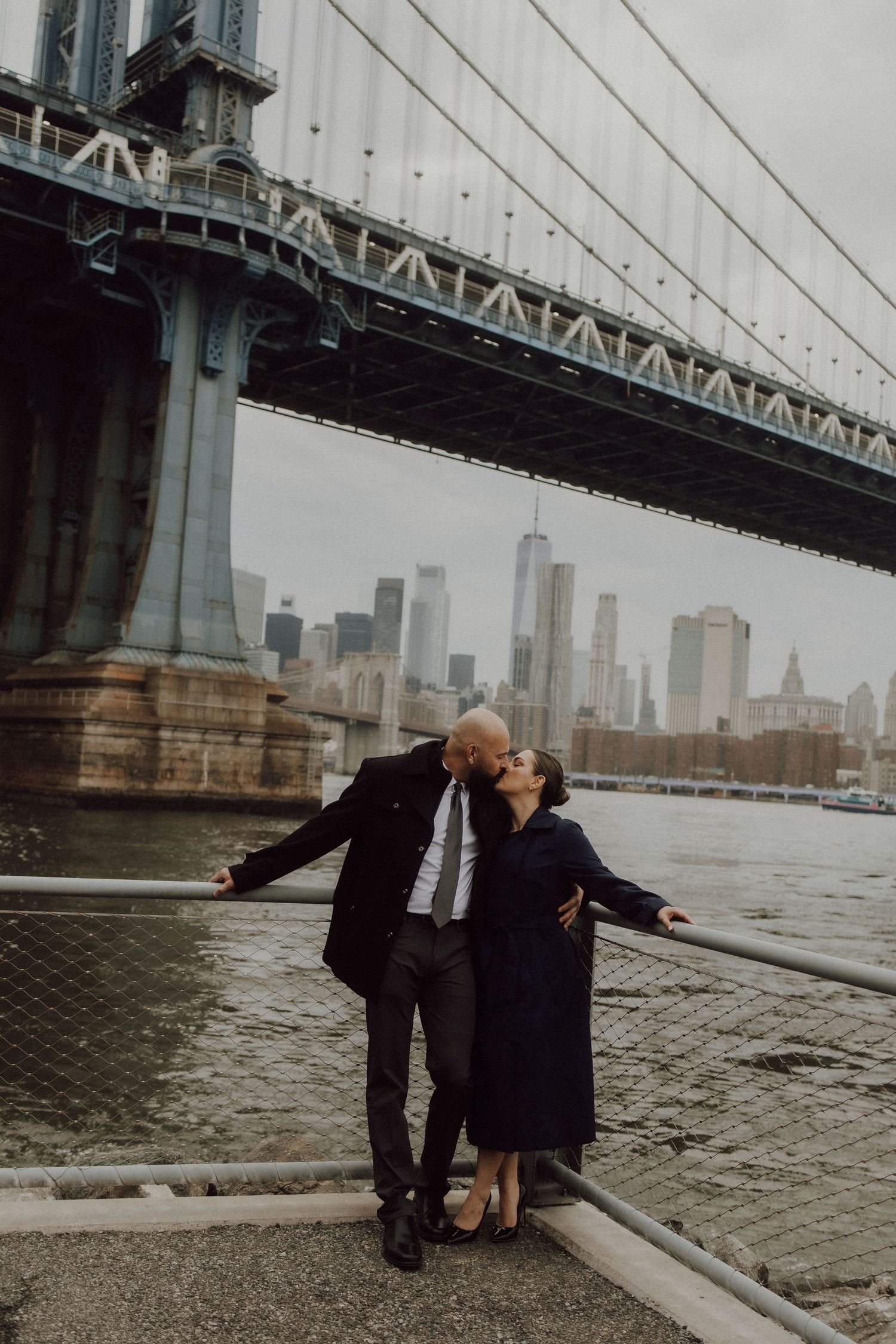 A couple shares a tender moment under the Manhattan Bridge with the city skyline in view.