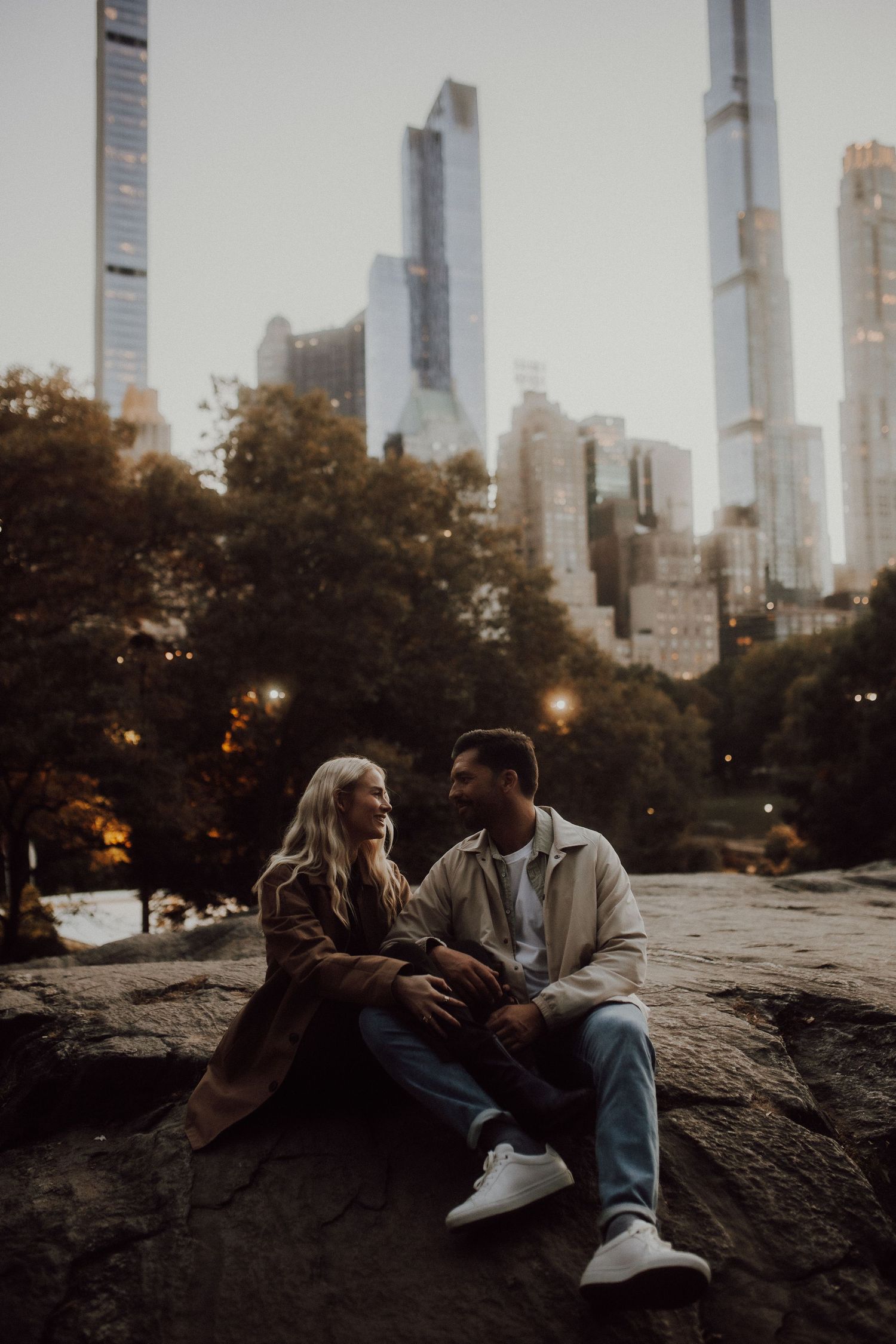 A couple relaxes on rocks in Central Park with Manhattan skyscrapers towering in the background.