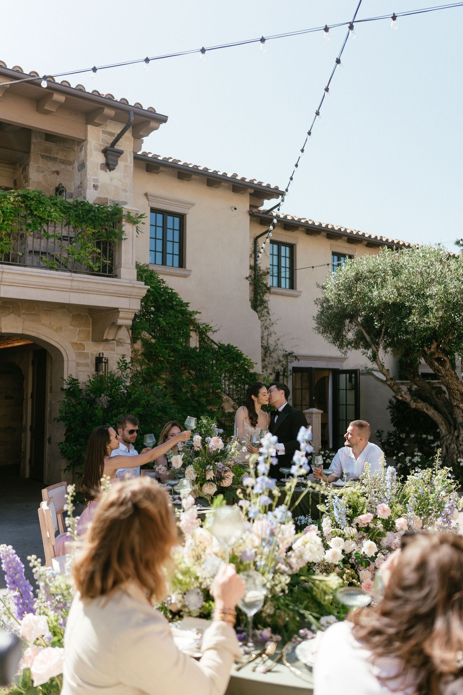 Outdoor wedding reception at a Tuscan-style villa with floral centerpieces, string lights, and guests toasting the couple.