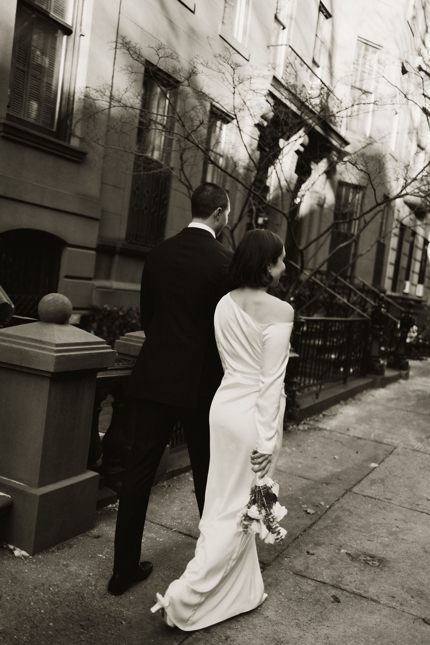 A bride in a white gown walks along a city sidewalk in black and white photography.