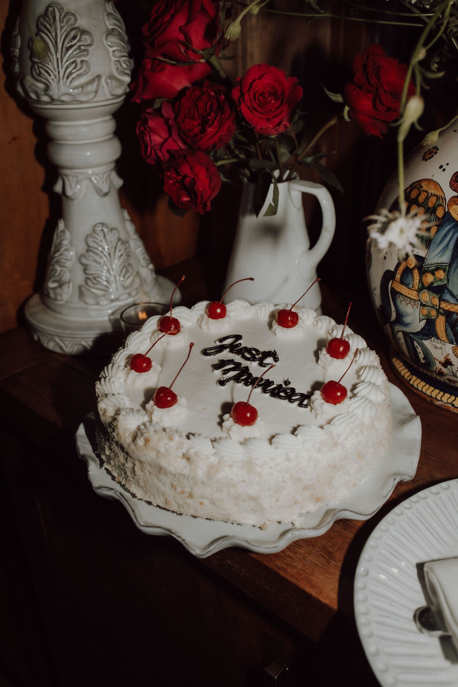 A white wedding cake decorated with red roses and cherries sits on an ornate display.