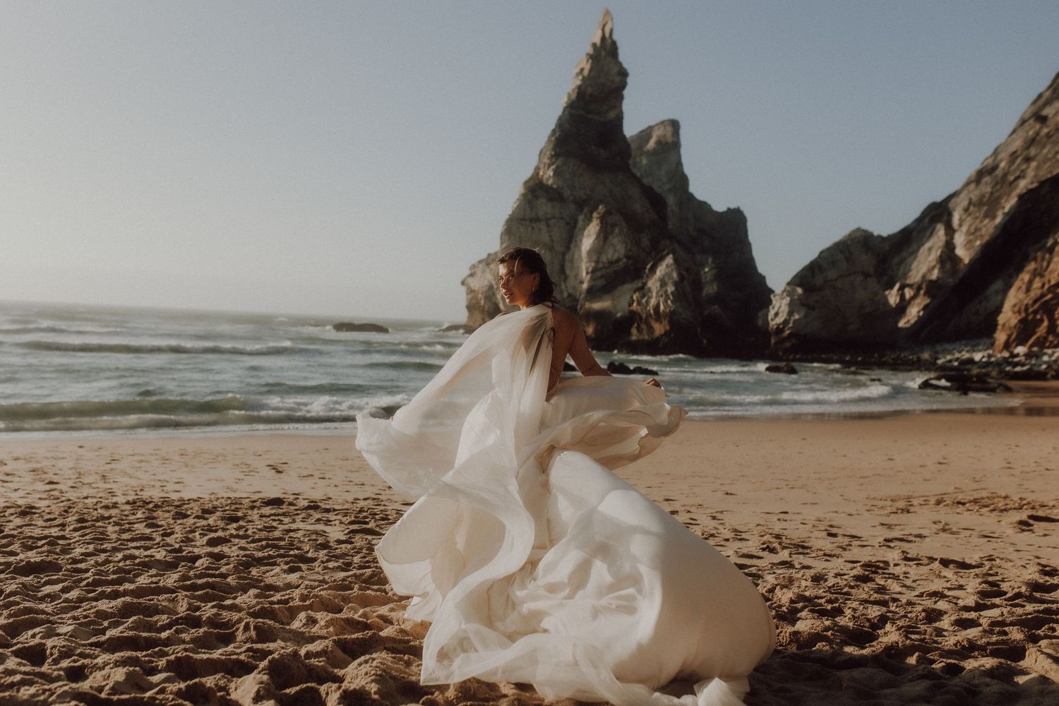 A flowing white wedding dress catches the breeze on a dramatic coastal beach with rocky cliffs.