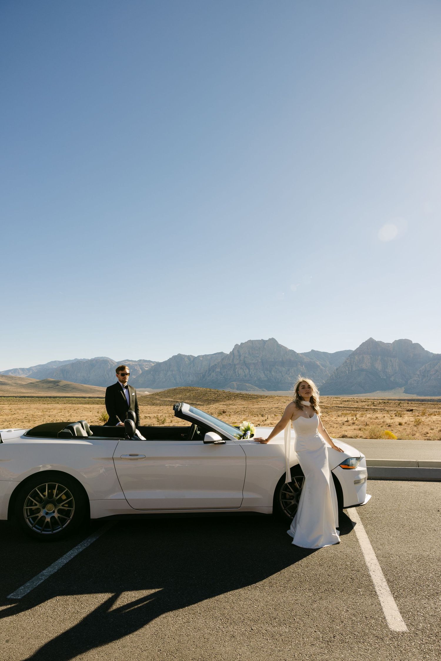 A desert wedding photoshoot featuring a white sports car on an empty road with mountain views.