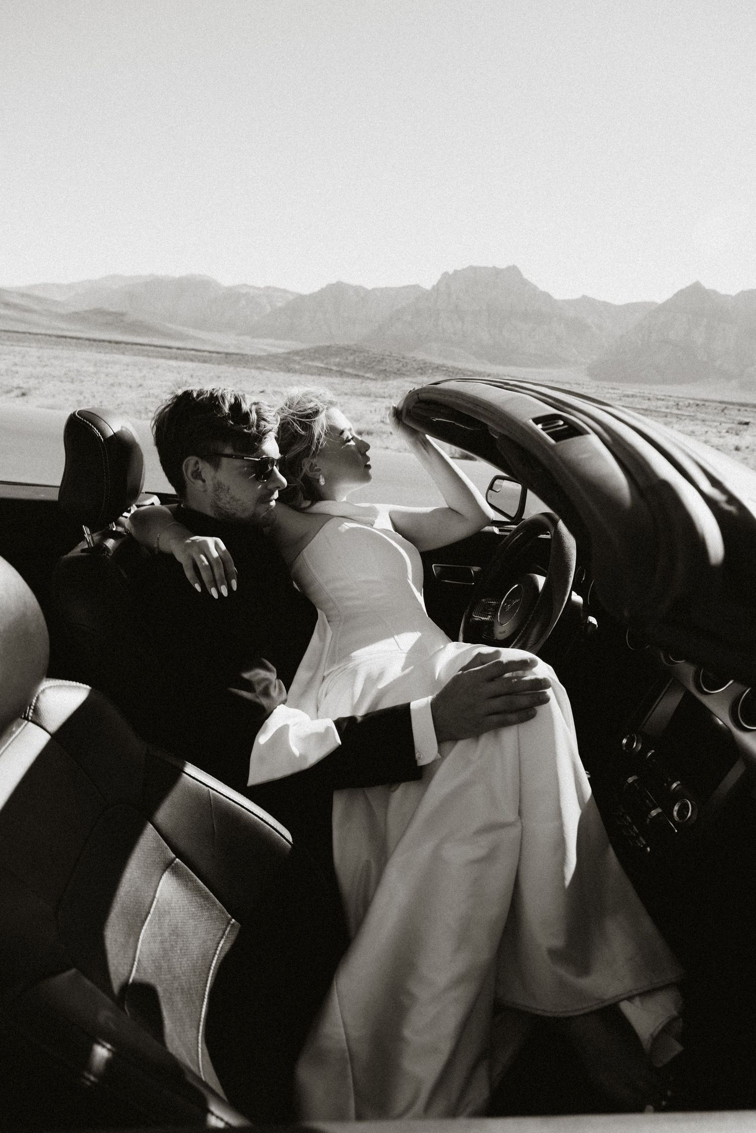 A black and white photo of people relaxing in a convertible car with mountains visible in the background.