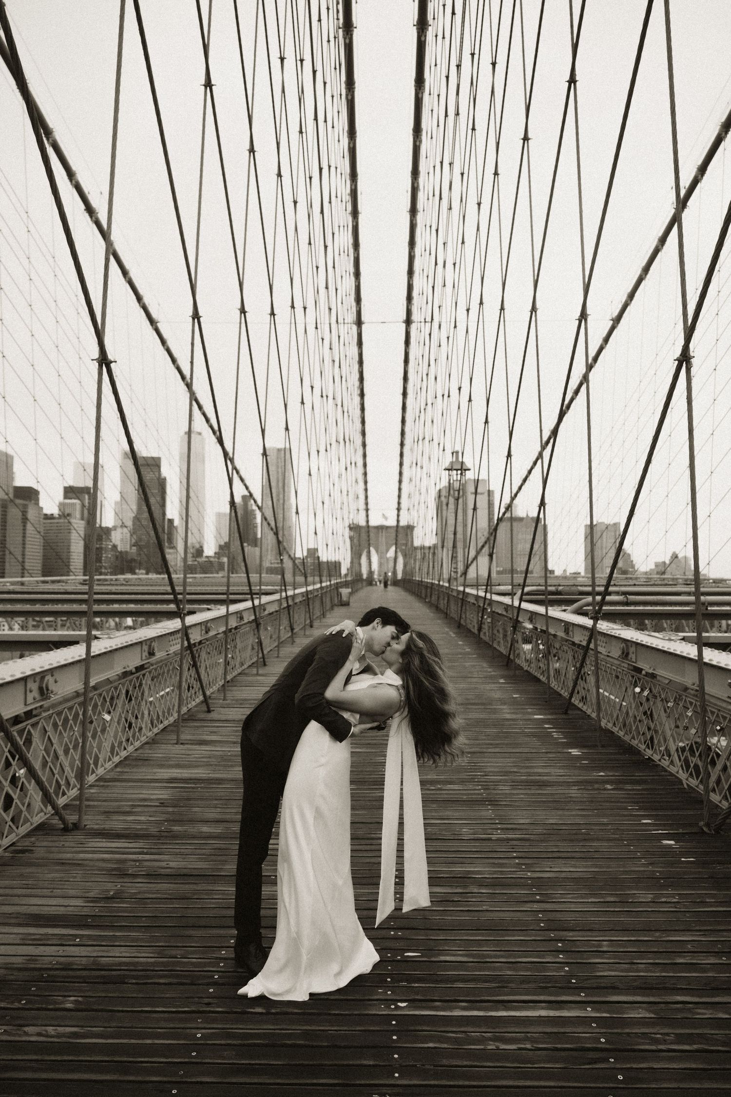 Black and white photo of a couple on the Brooklyn Bridge with city skyline in background.