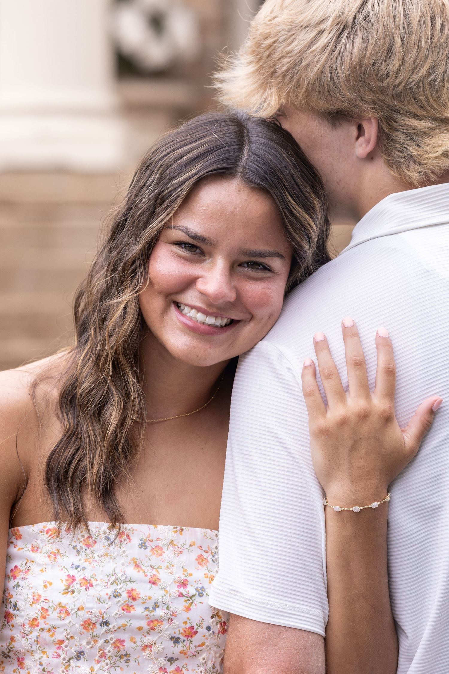 A smiling young woman in a floral dress rests her head on a man's shoulder, showing an engagement ring.
