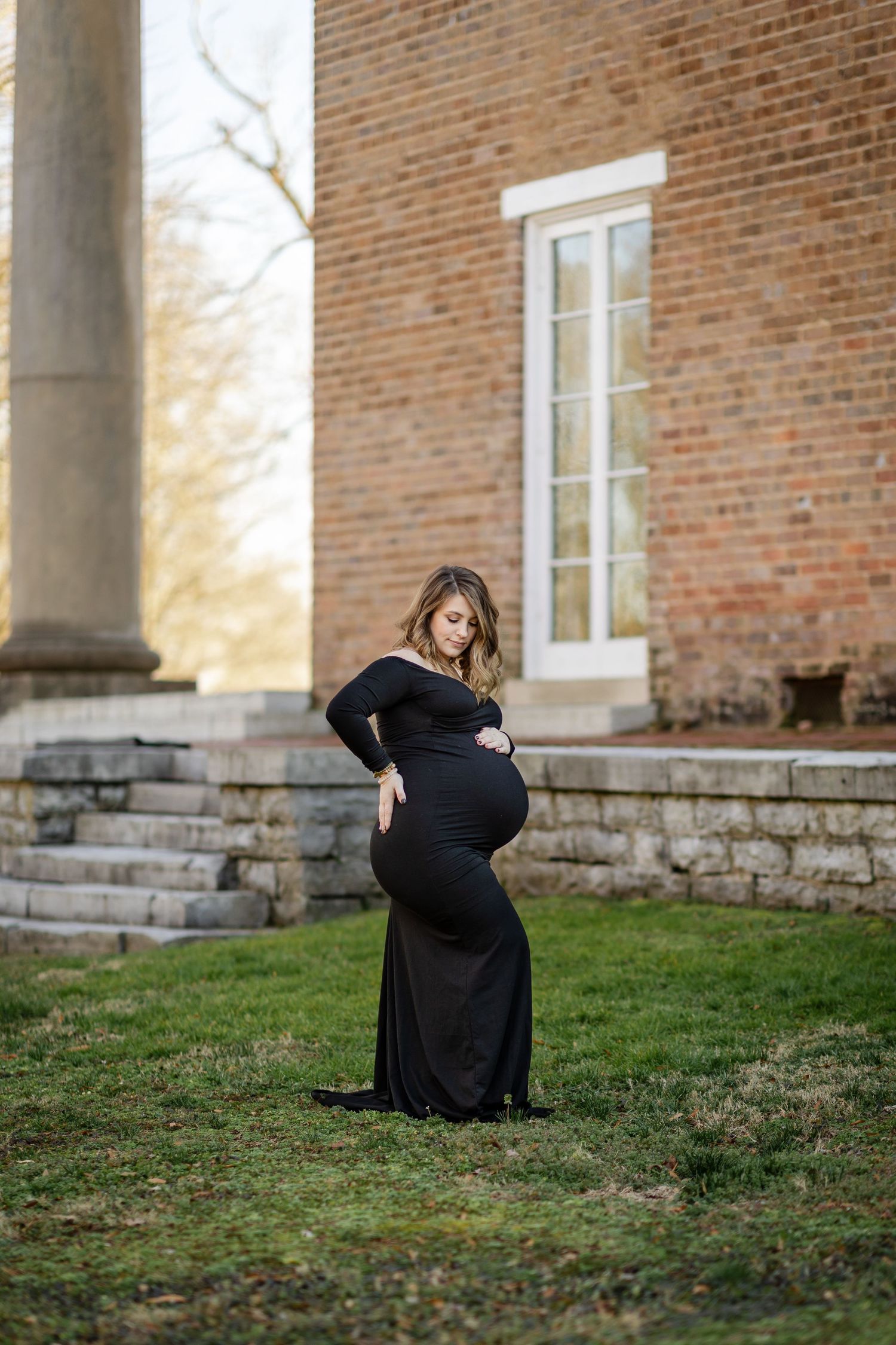 Pregnant woman in black gown cradling baby bump outdoors near brick building with columns and stone steps.