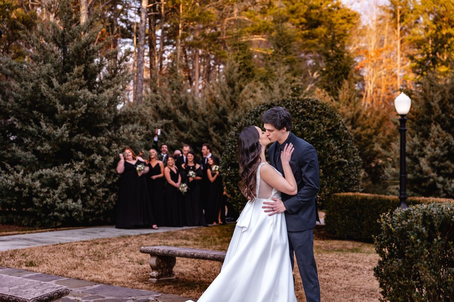 Bride and groom sharing a first kiss outdoors with bridesmaids in black dresses standing behind them.