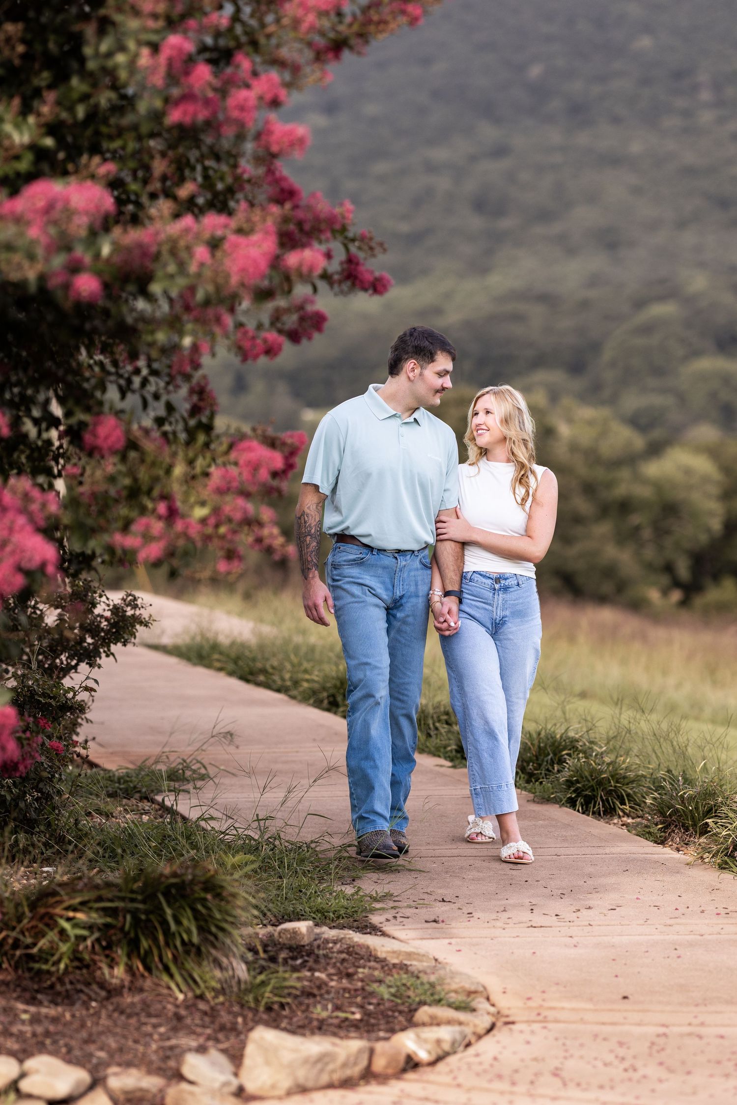 Couple walking hand-in-hand on a scenic path with pink blooming trees and green hills in the background.