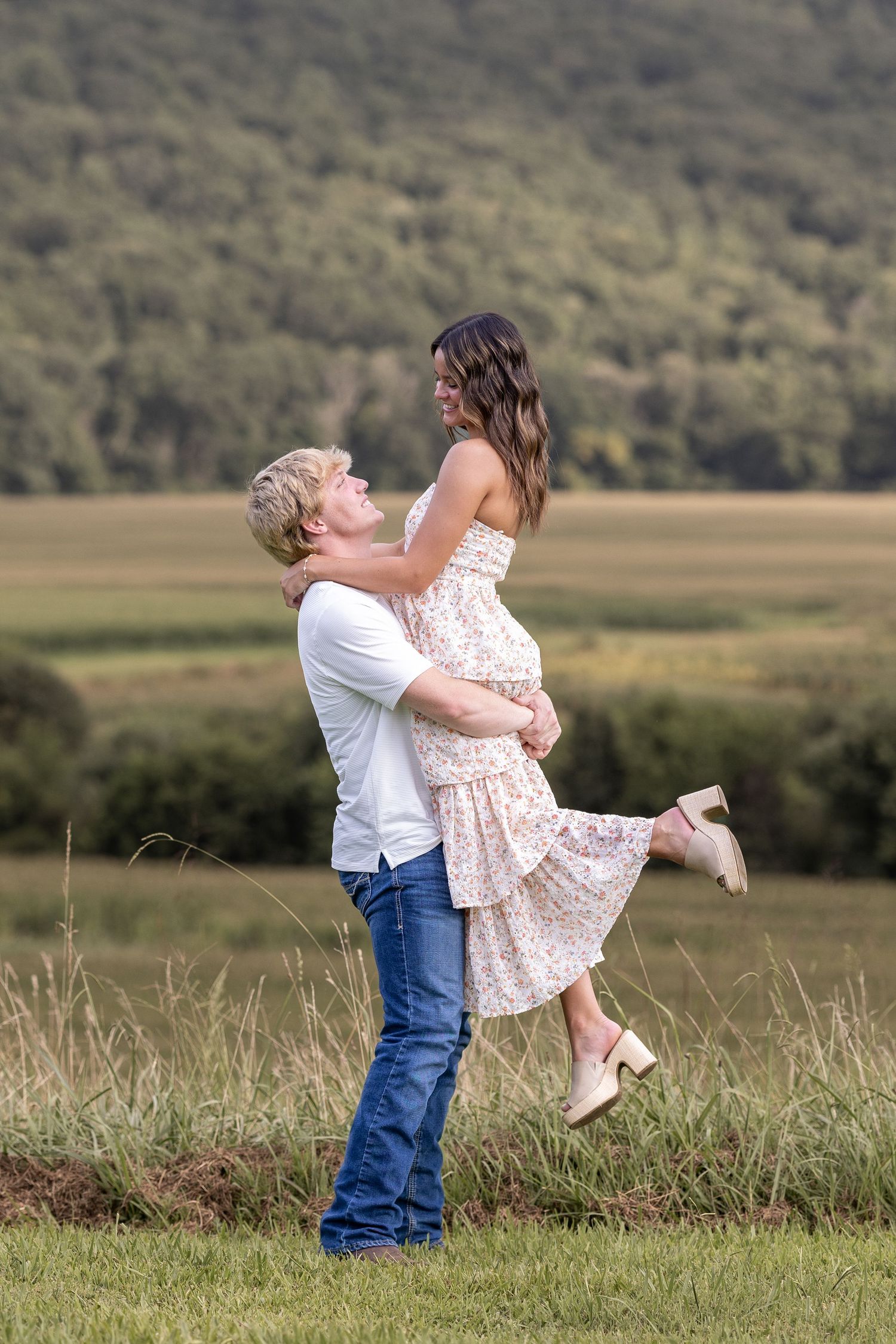 A man lifts a woman in a floral dress in a scenic green field, sharing a romantic moment outdoors.