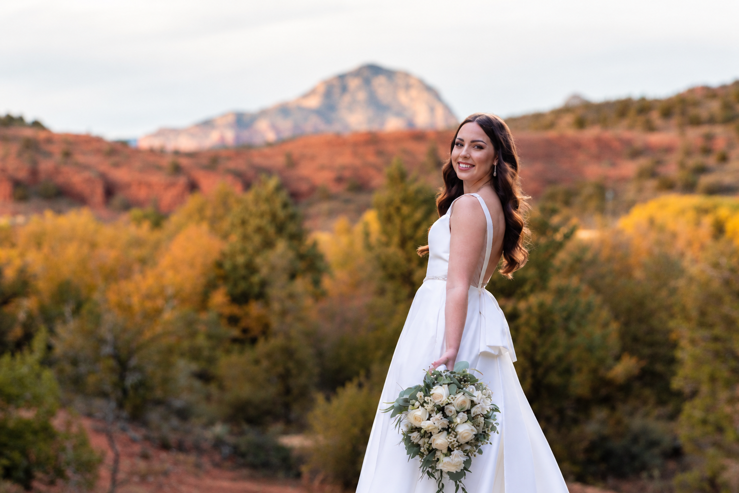 Bride in white gown holding bouquet poses before red rock mountains and autumn foliage in Sedona, AZ.