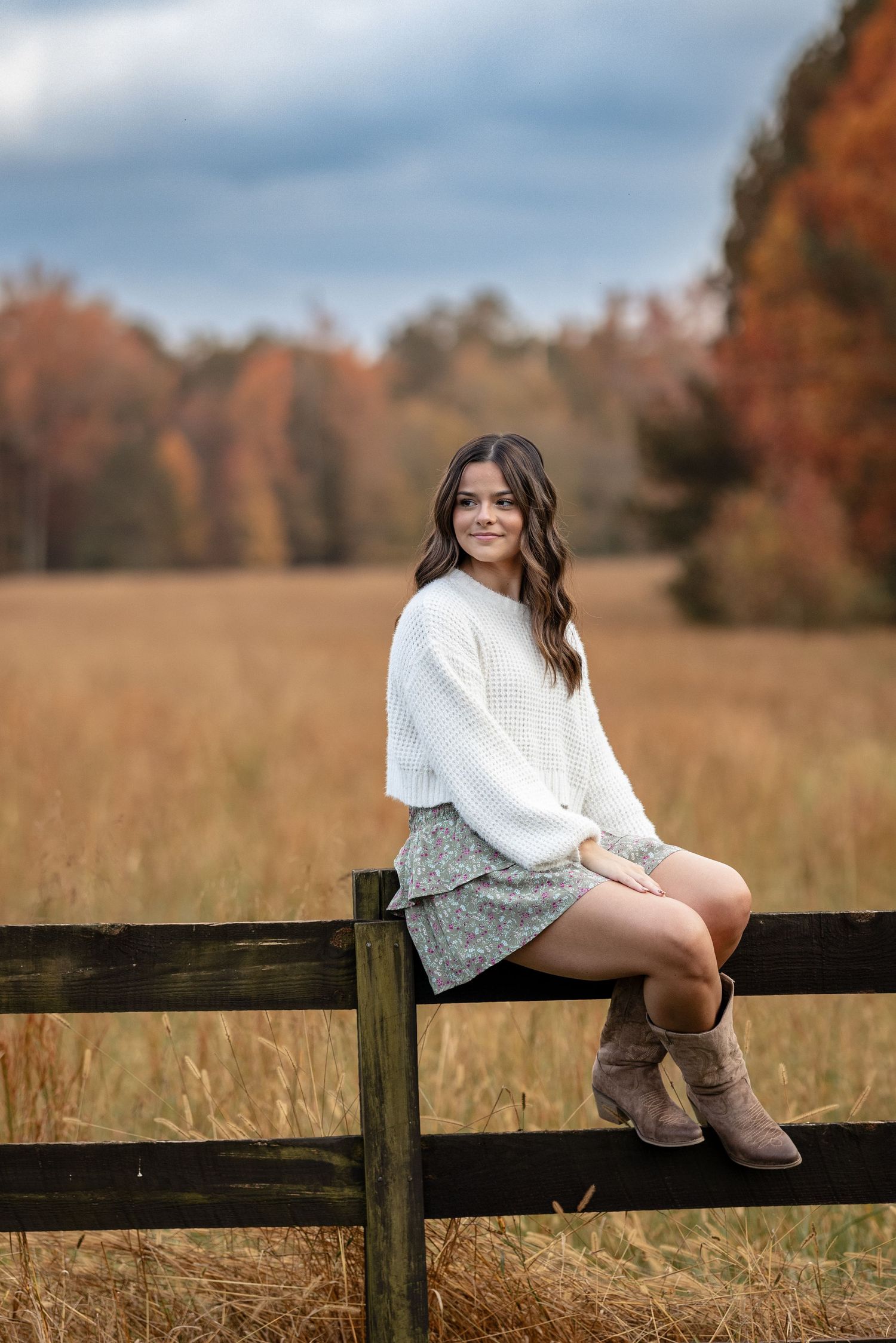 Young woman in white sweater and floral skirt sits on wooden fence in autumn field with colorful fall foliage.
