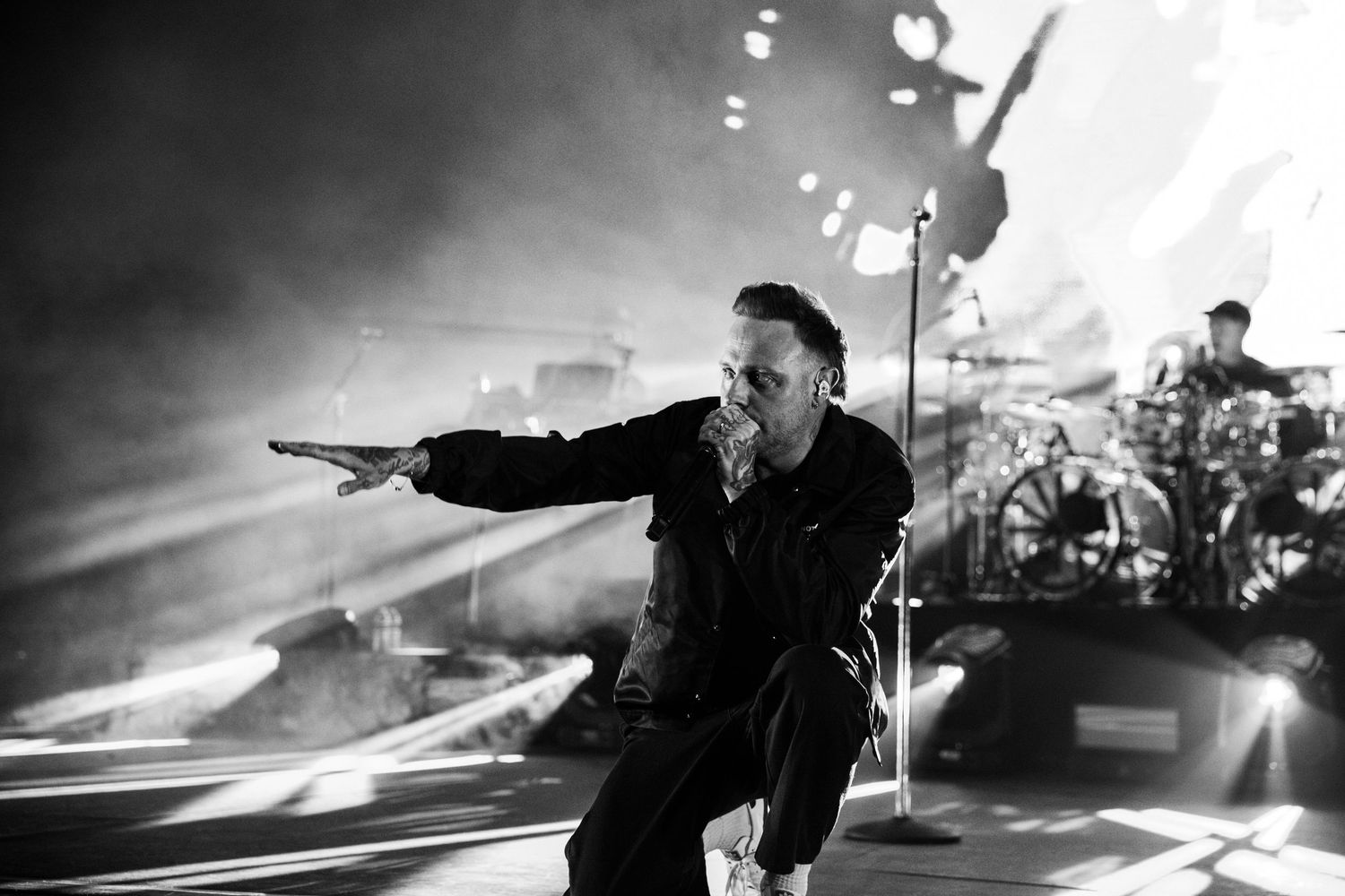 Black and white concert photo of a performer pointing outward on stage with dramatic lighting and band members behind.