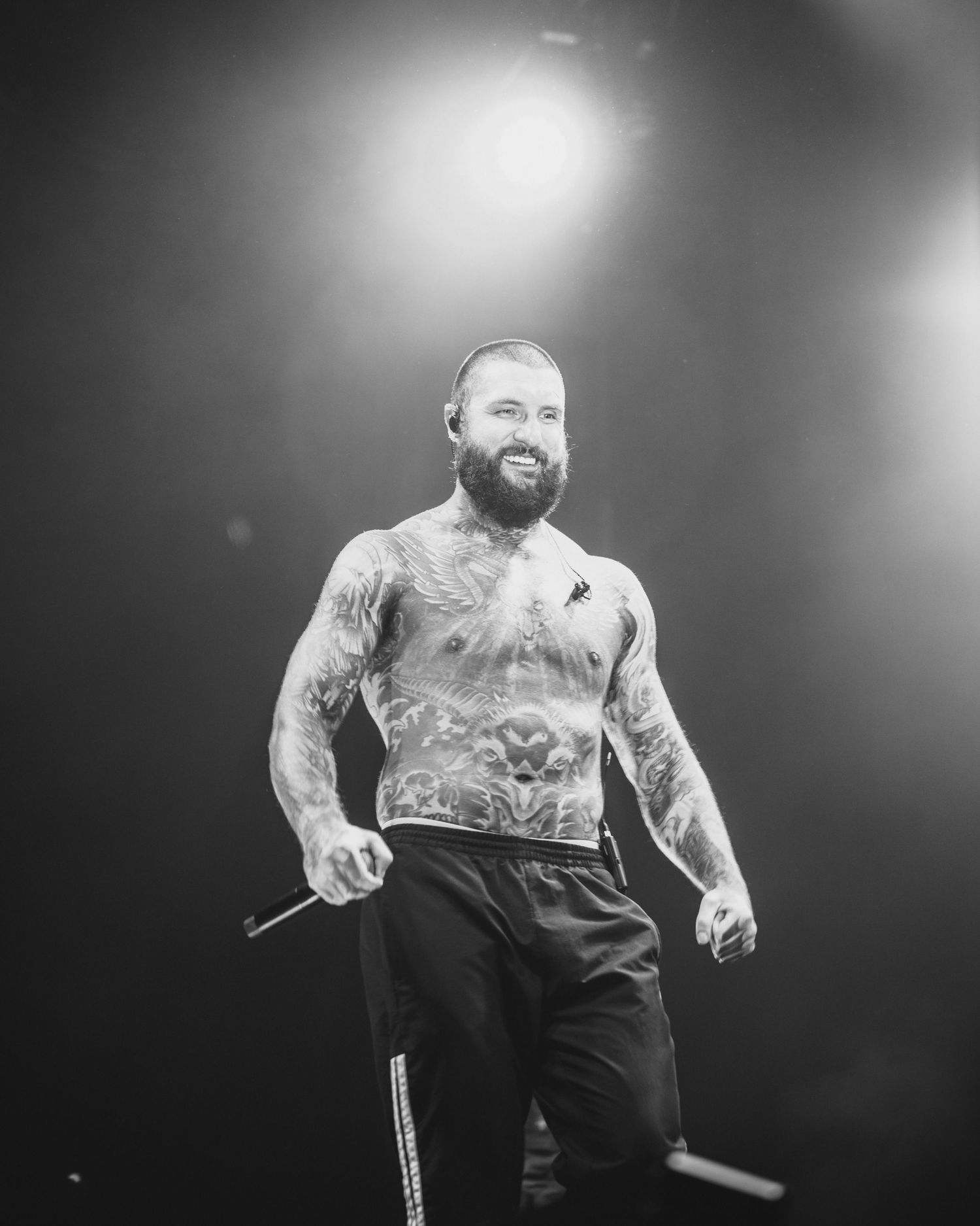 Tattooed shirtless male performer smiling on stage holding a microphone in black and white.