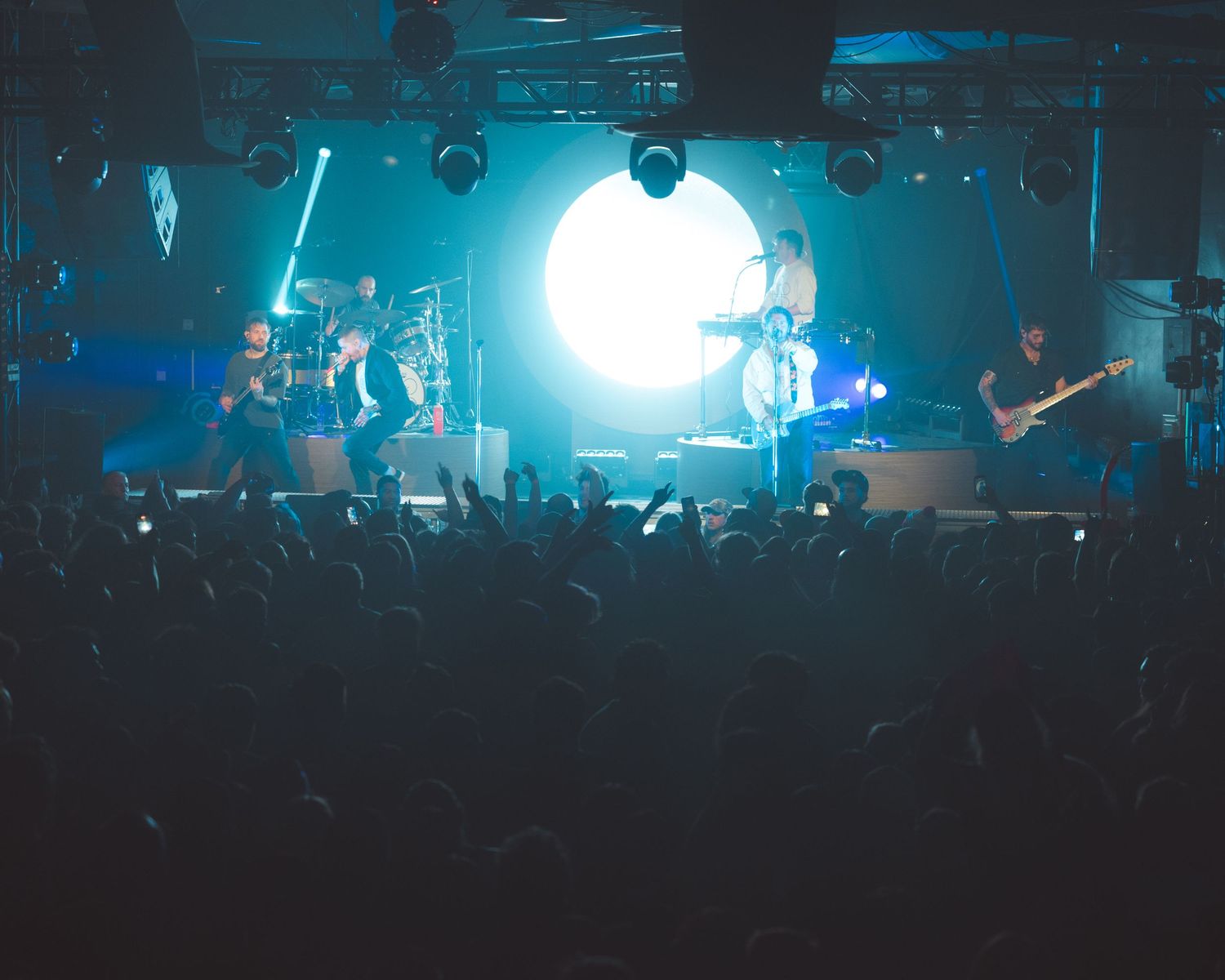A rock band performs on stage before a large crowd with dramatic white spotlight and blue concert lighting.