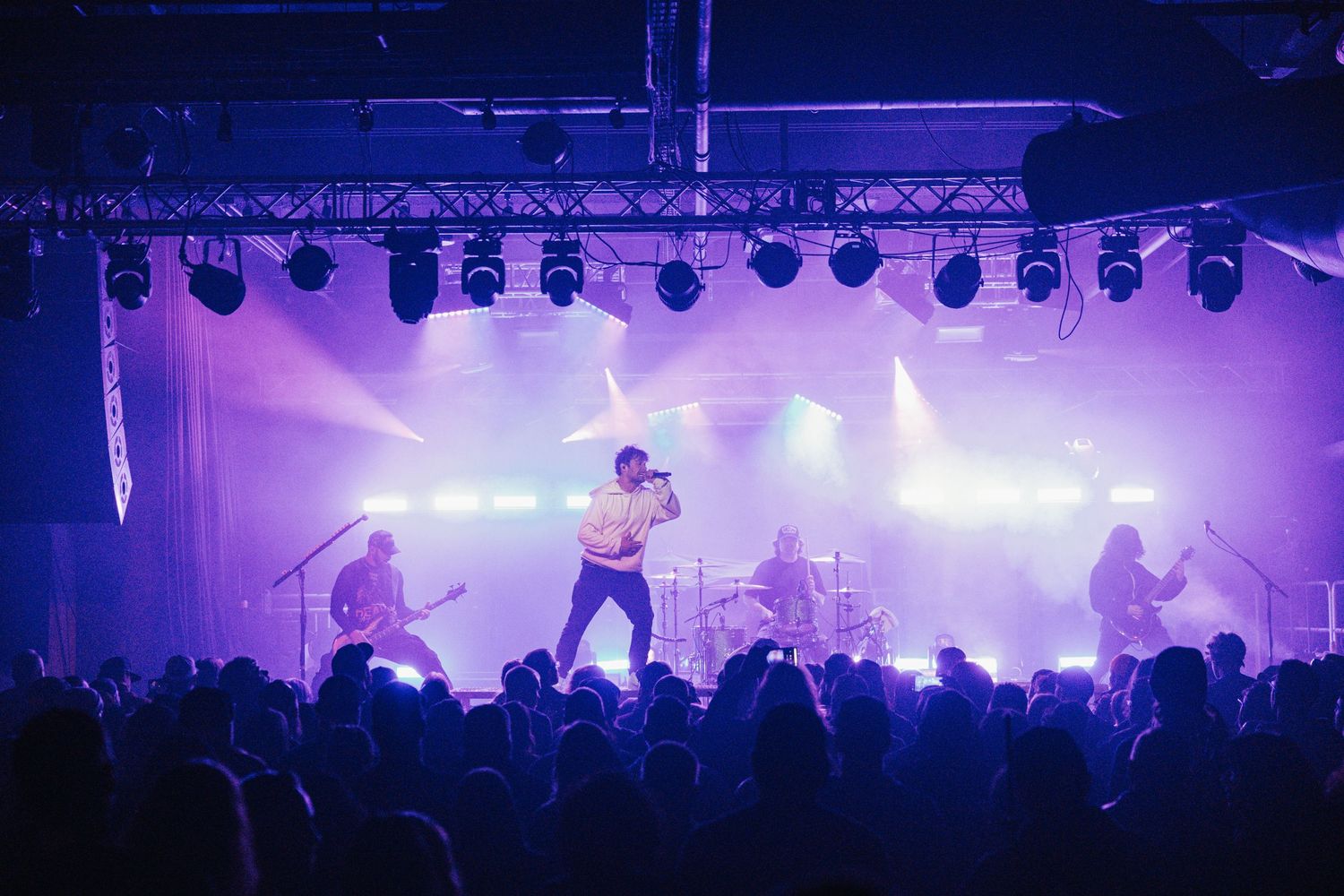 A rock band performs on stage with dramatic purple lighting and fog effects as silhouetted crowd watches from below.