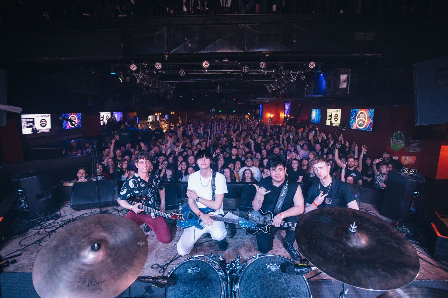 Wide angle shot of a packed concert venue showing crowd and band performing on stage with drum kit visible.