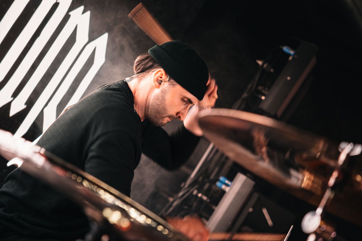 A focused male drummer wearing a black beanie plays cymbals in a dimly lit music venue.