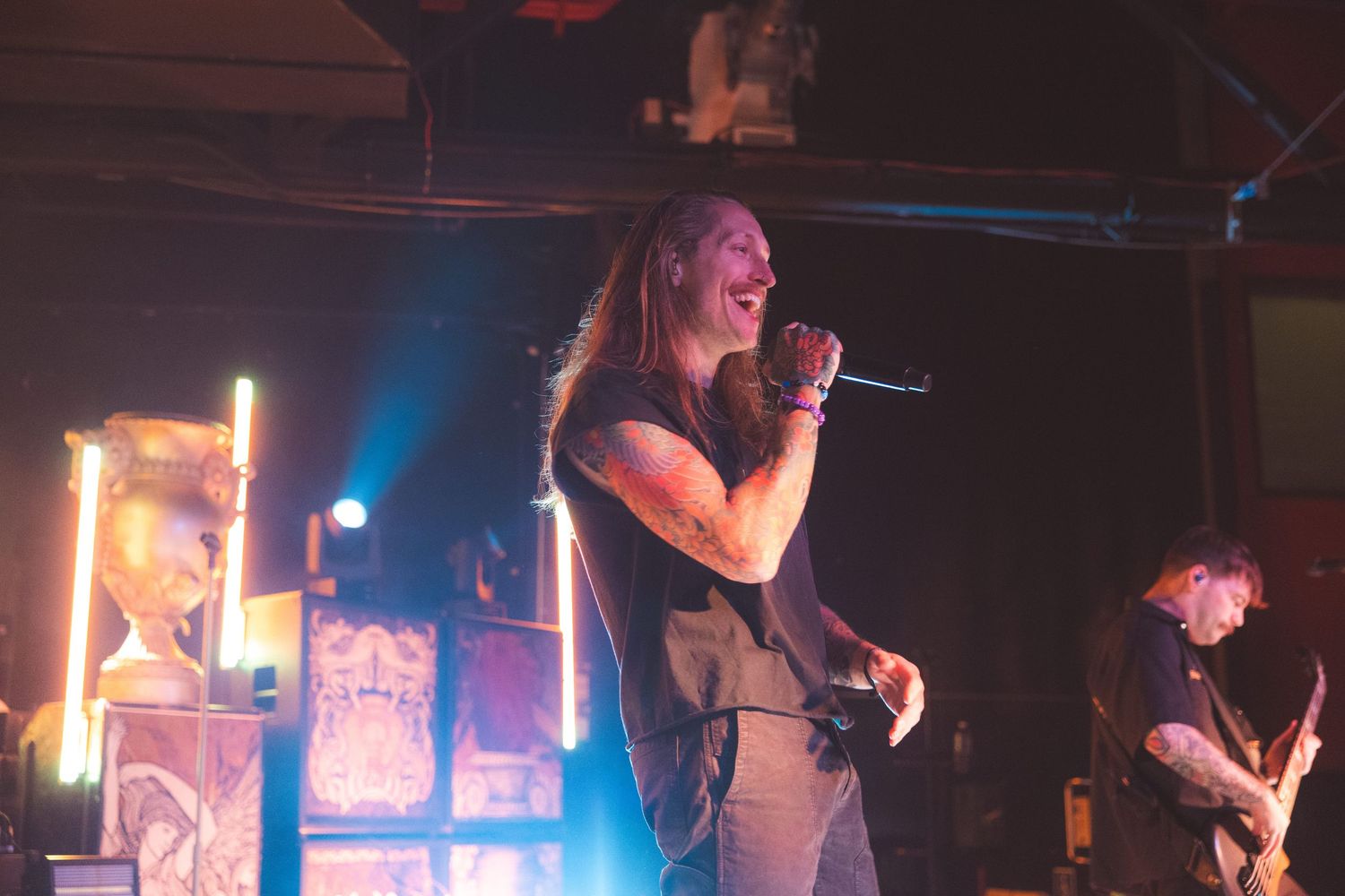 A performer sings on stage with colorful blue lighting and amplifier stacks visible in the background.