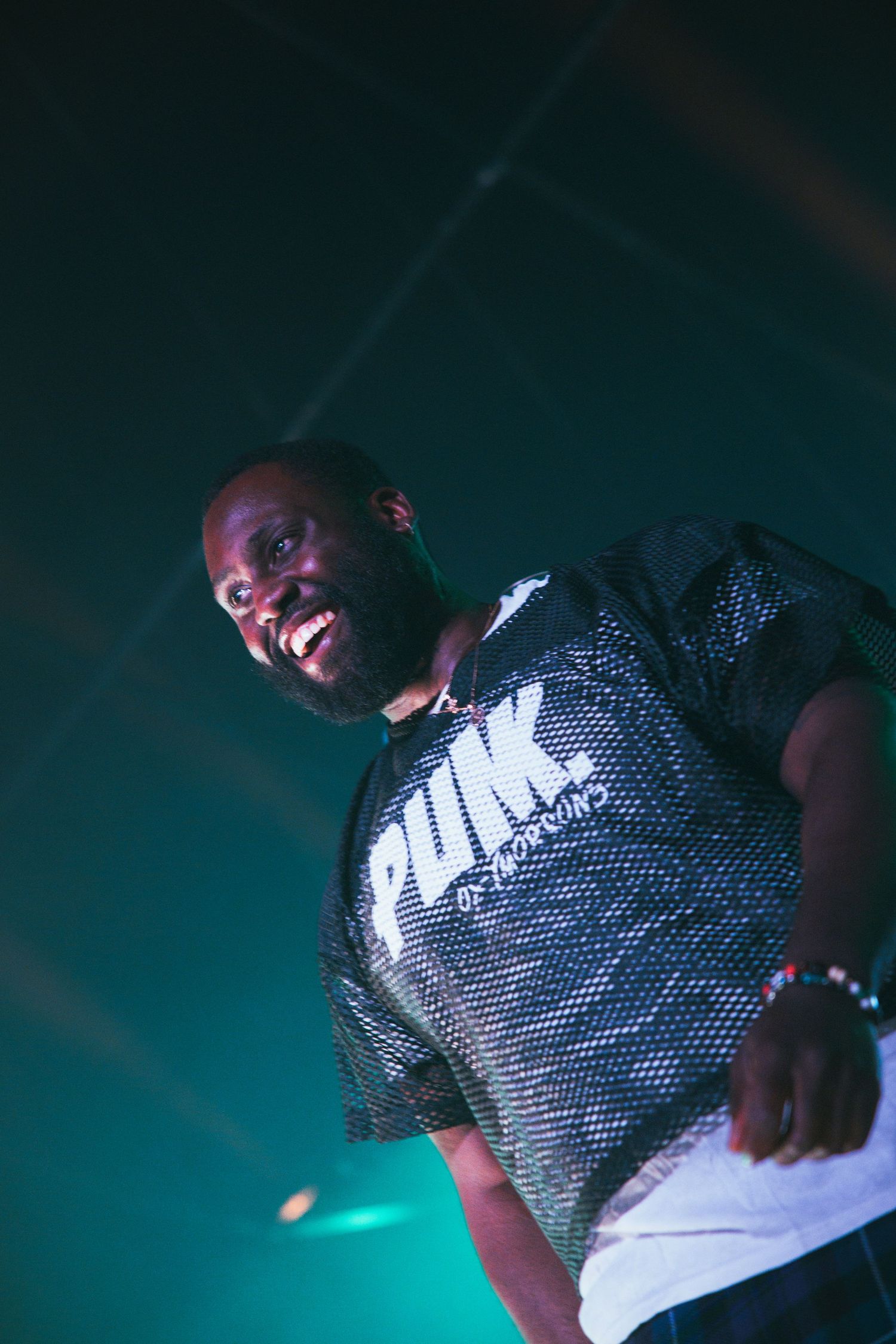 Performer wearing shiny metallic shirt smiles broadly while performing on stage with teal lighting in background.