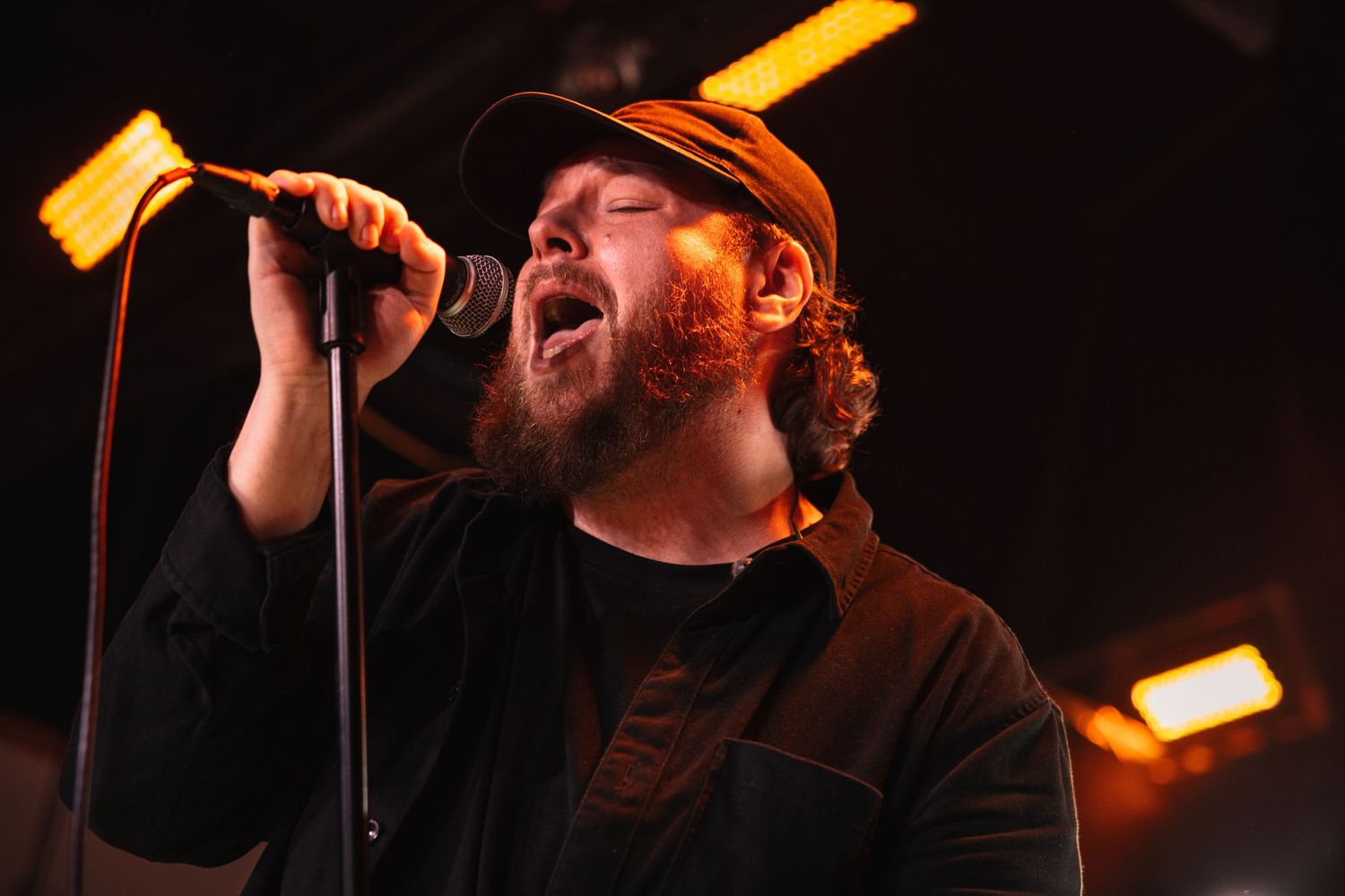 Bearded male vocalist singing passionately into a microphone on stage under warm amber lights.