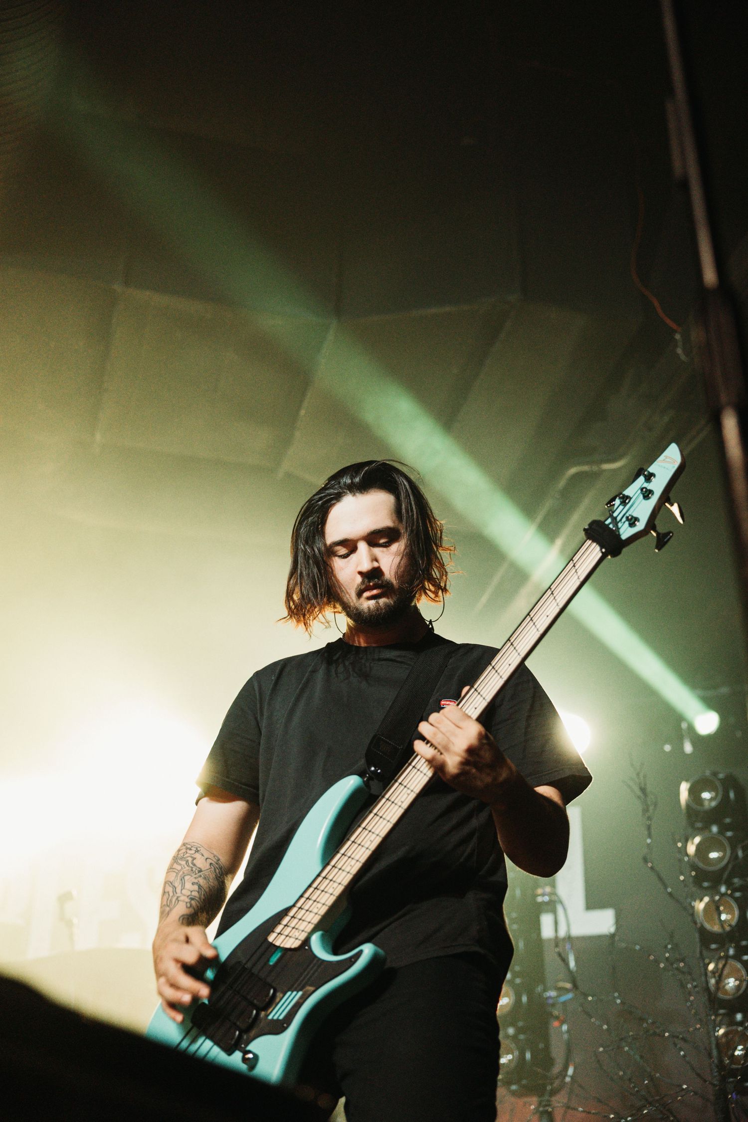 Rock musician performs on stage with a black electric bass guitar under dramatic green lighting.
