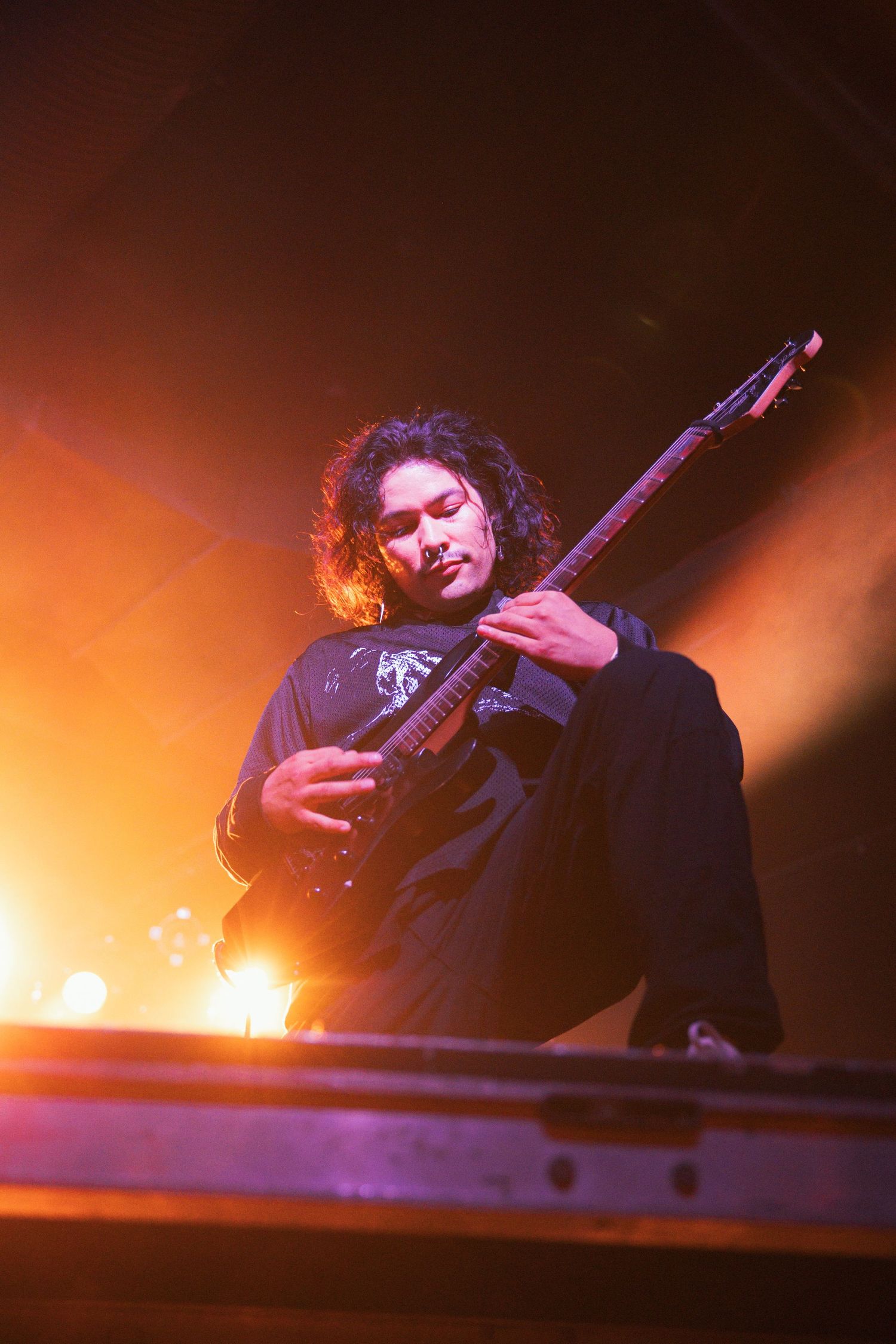 Musician plays bass guitar on stage with dramatic orange lighting and smoke effects in the background.
