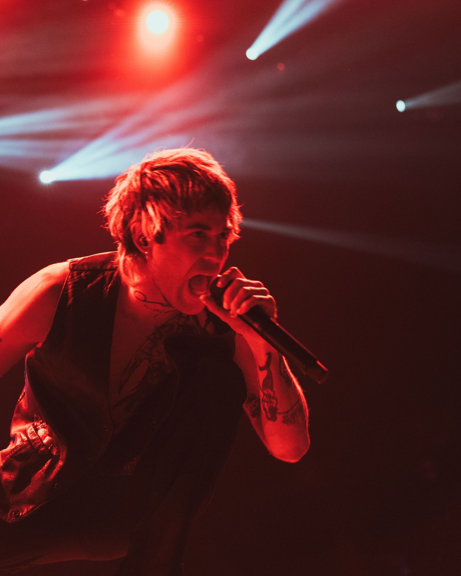 Tattooed singer performing on stage under dramatic red lighting with blue stage beams at a live concert.