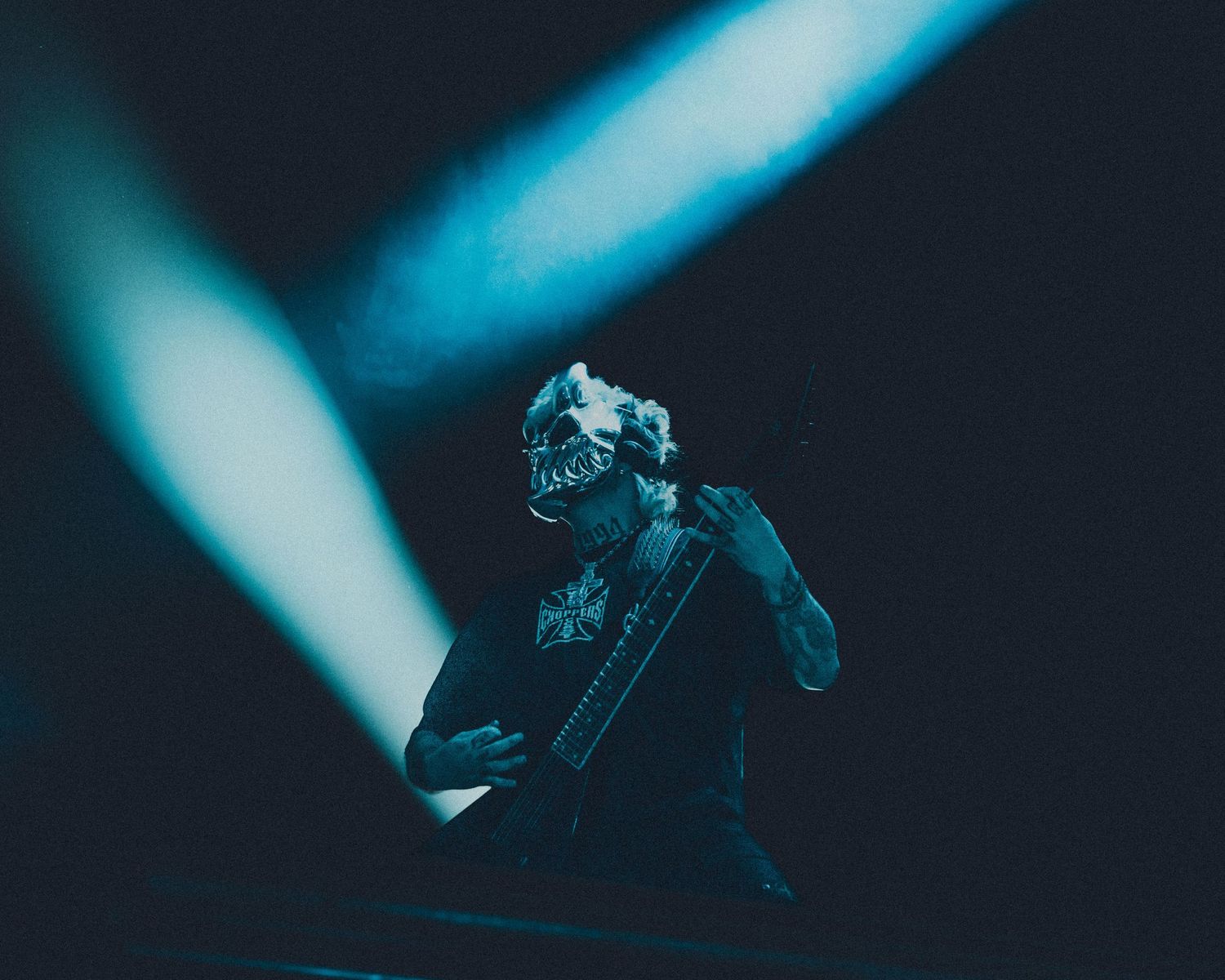 Masked performer playing keyboard on dark stage with dramatic blue spotlight beams during live concert.