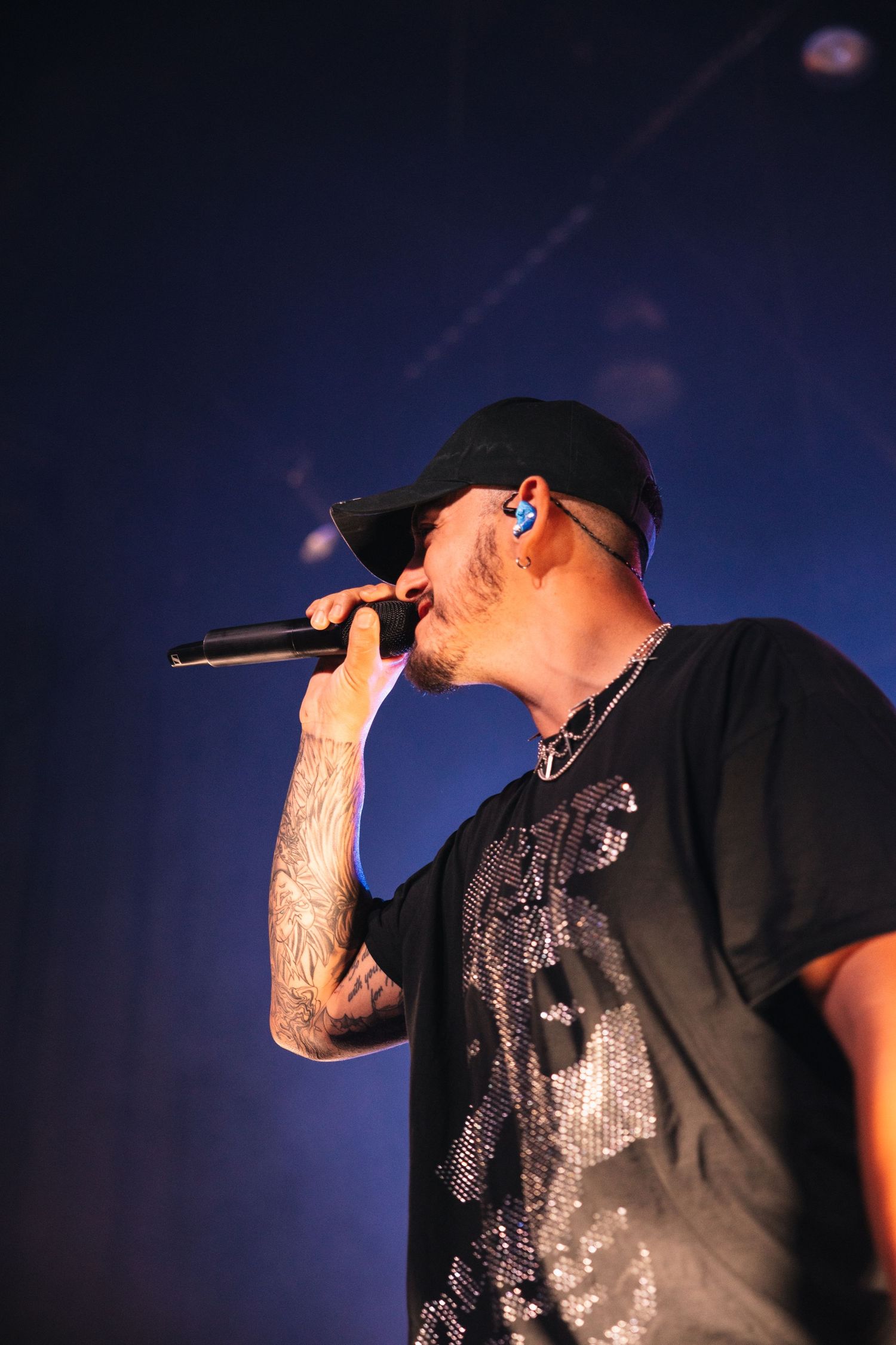 A performer on stage in a black t-shirt and baseball cap sings into a microphone under dark blue lighting.