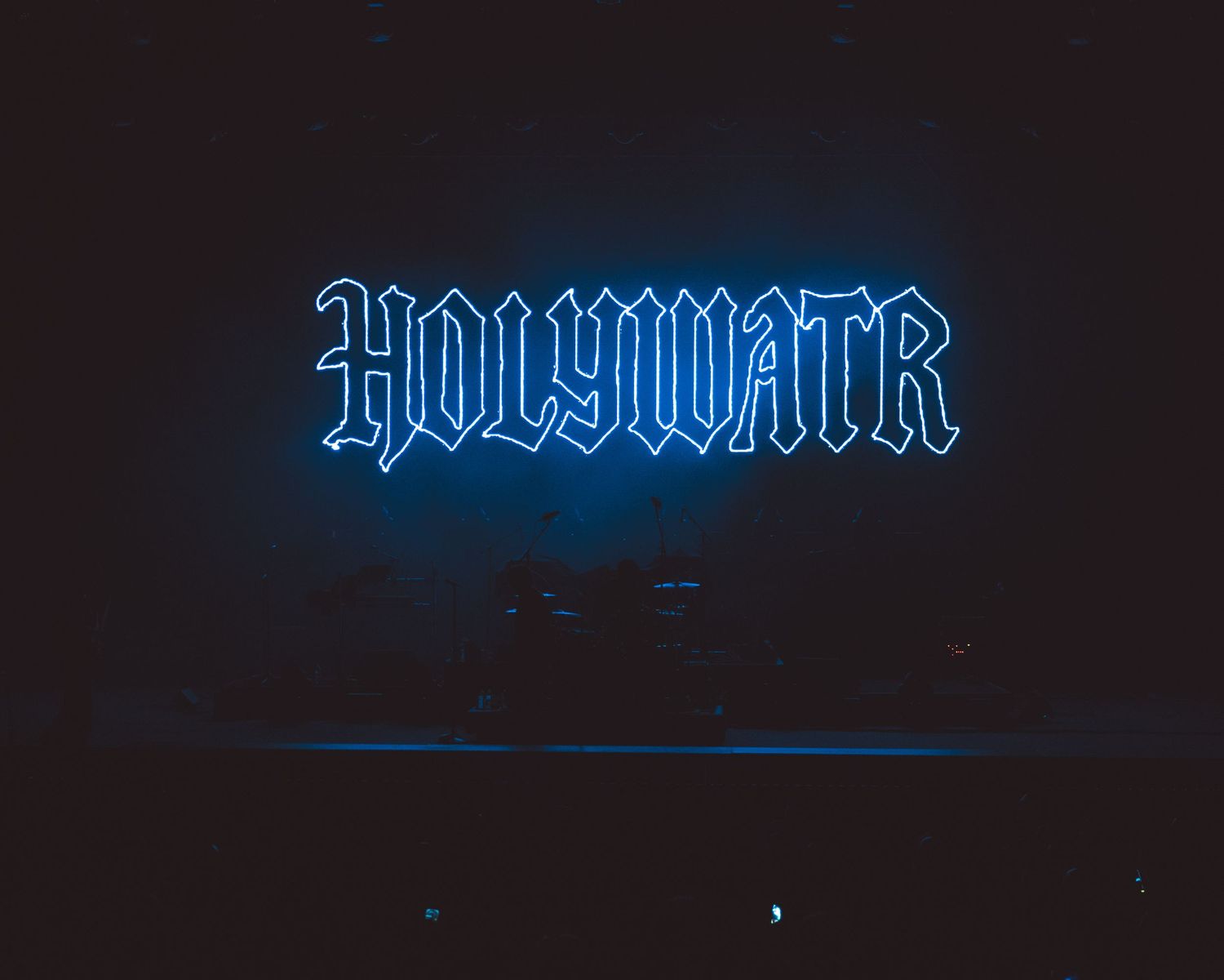 Glowing blue neon sign reading HOLYWATR in gothic lettering against a dark background at a concert venue.