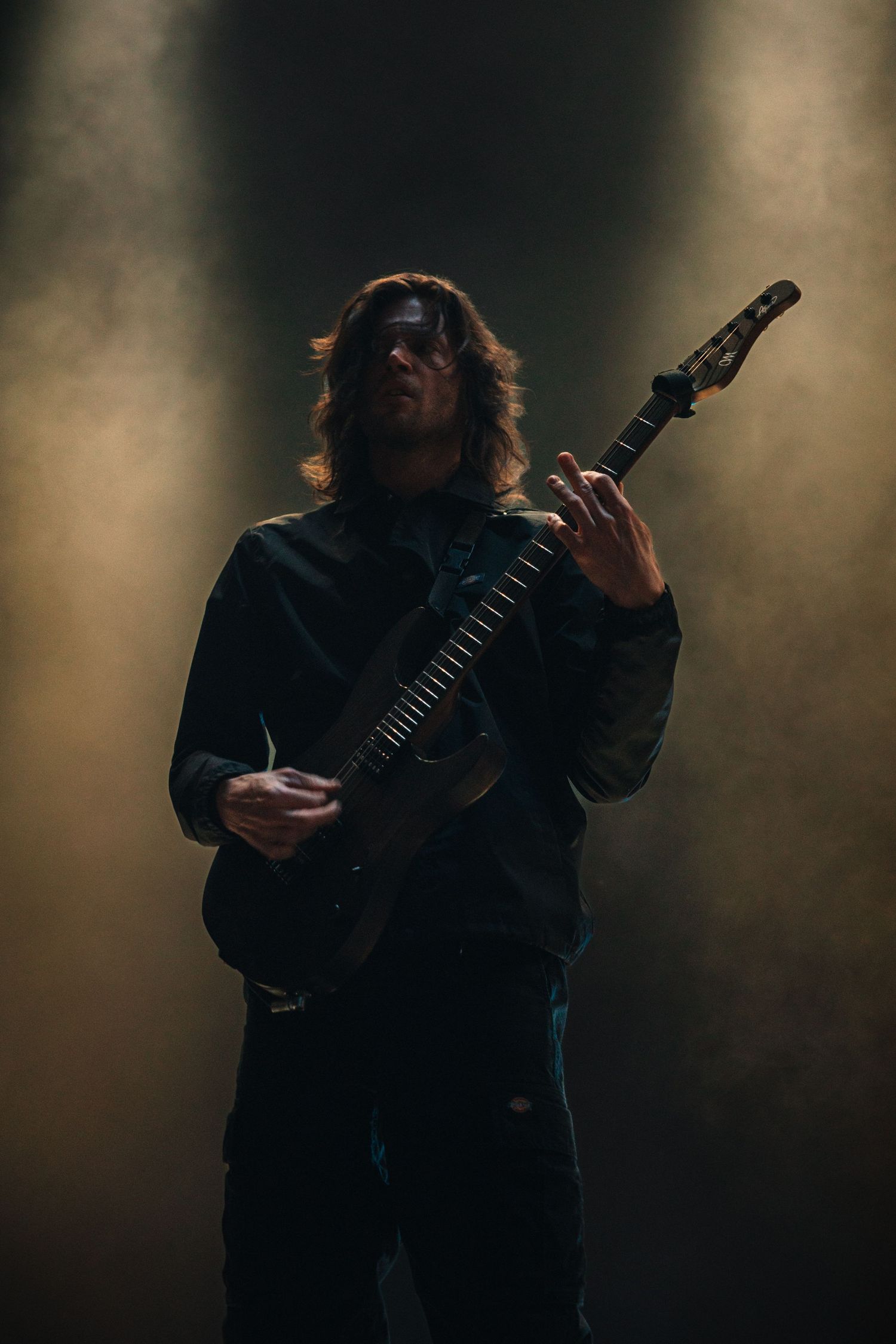 Long-haired guitarist performs on smoky stage with dramatic lighting, playing electric guitar in dark concert setting.