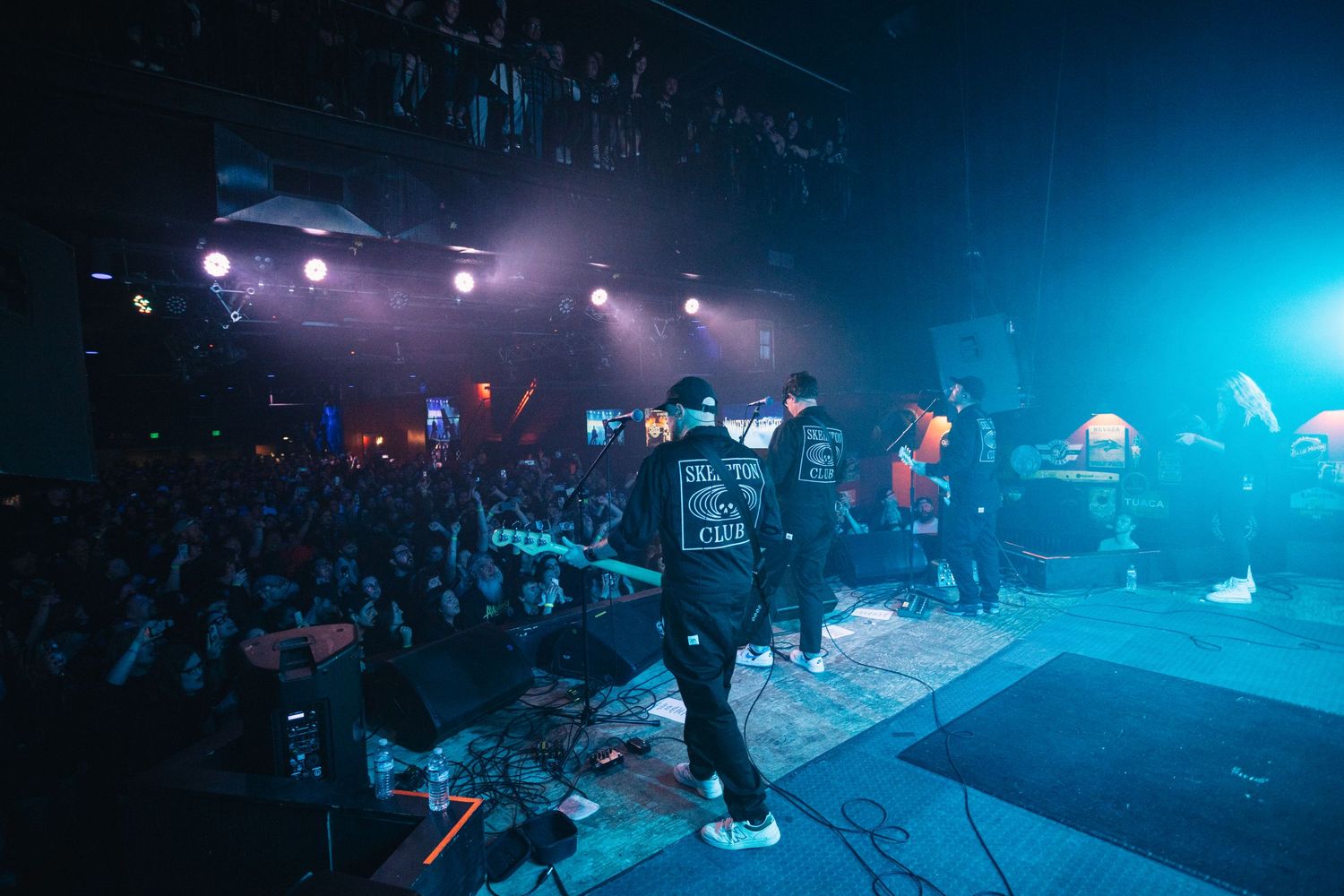 Performers on stage with energetic crowd at dimly lit concert venue with blue lighting effects.