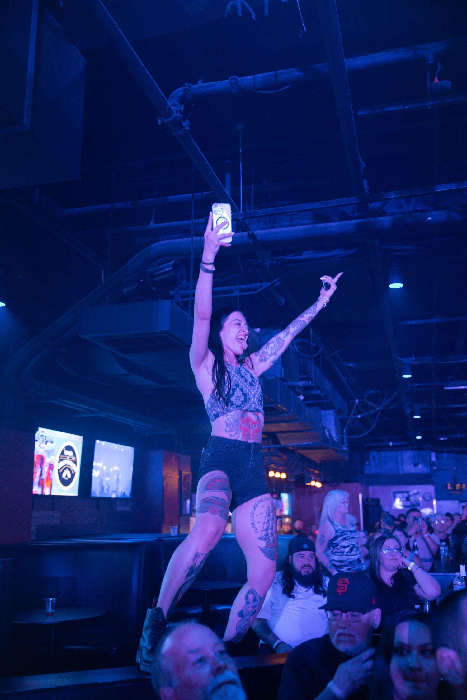 A person at a concert stands on a crowded dance floor with arms raised under blue lighting in a nightclub atmosphere.