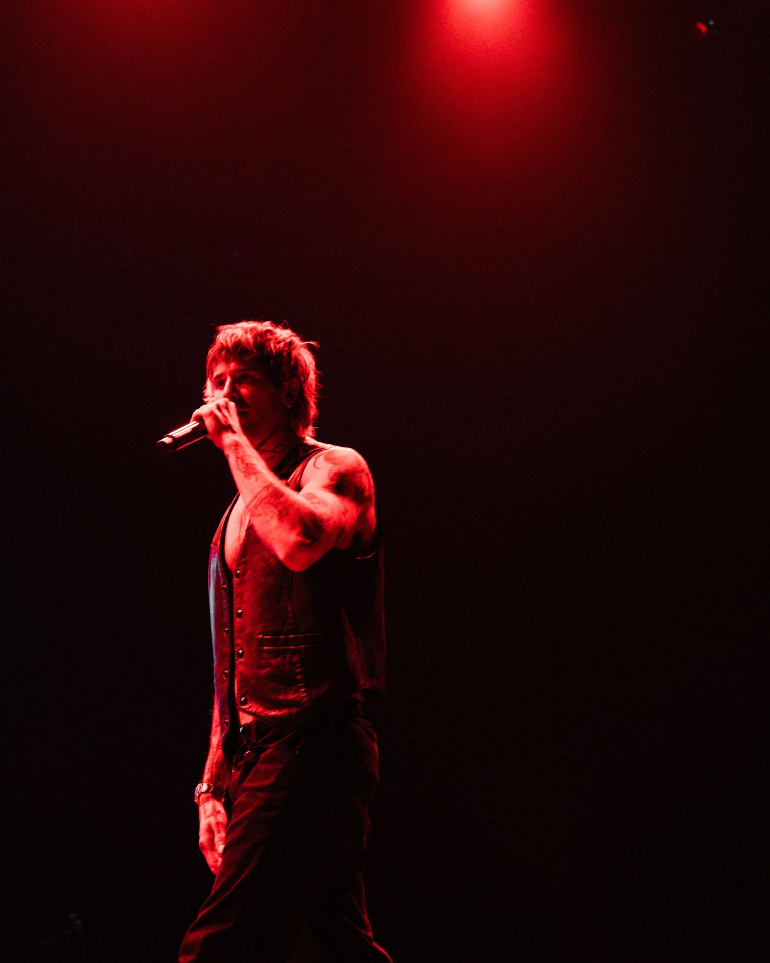 A shirtless male singer performs on stage under dramatic red lighting, holding a microphone close to his mouth.