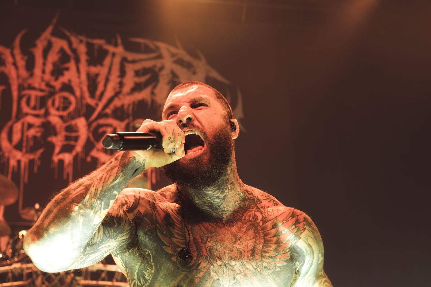 Heavily tattooed metal vocalist screaming into microphone on stage at a live concert performance.