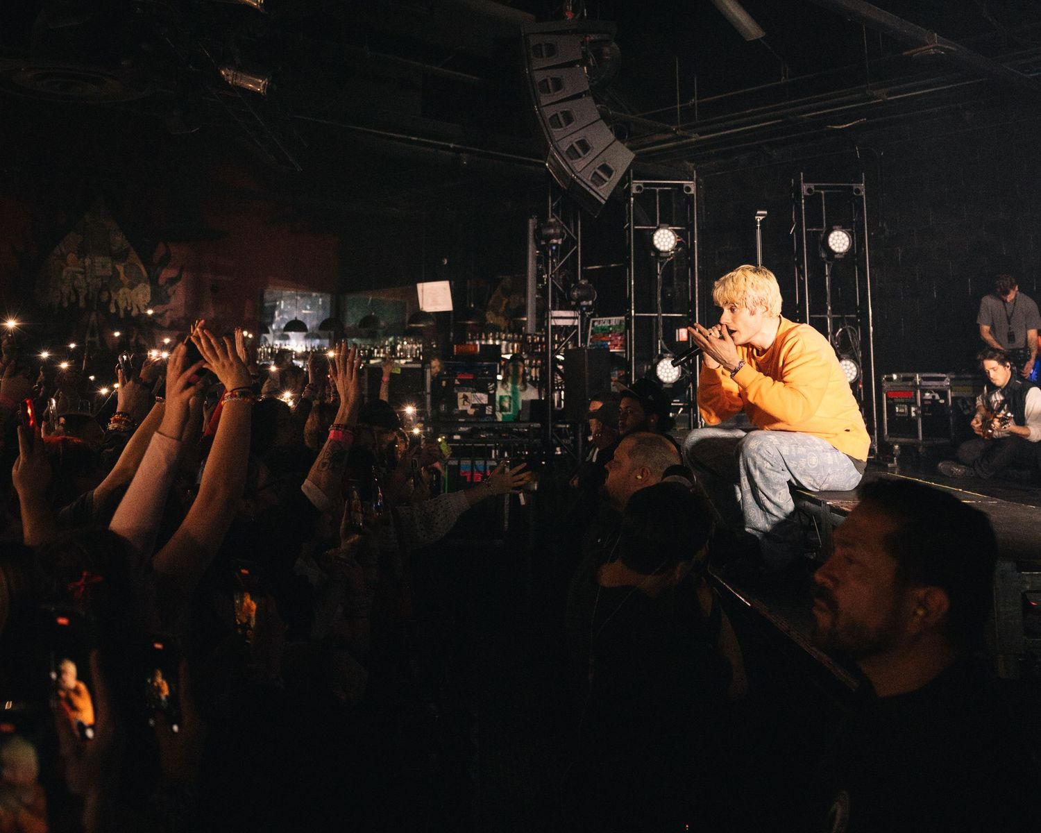 Concert crowd watching performers on stage in a dark venue with dramatic lighting and raised hands in silhouette.