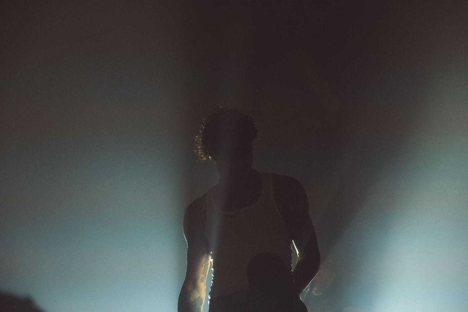 A dark silhouette of a person stands against a dramatic blue and teal backlit background creating an atmospheric scene.