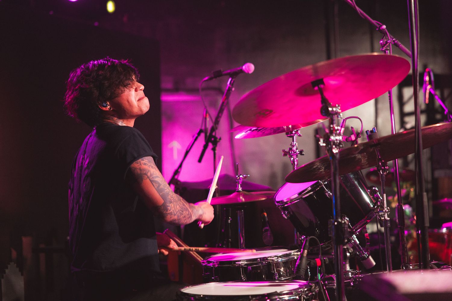 A drummer performs on stage with dramatic pink and purple lighting during a live music performance.