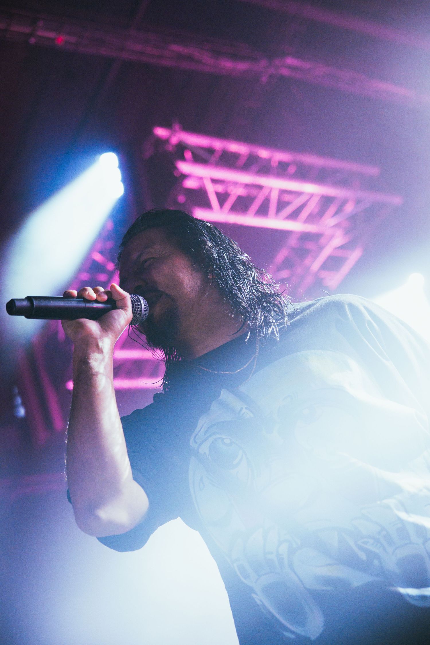 A performer sings into a microphone under dramatic stage lighting with blue and pink hues at a concert venue.