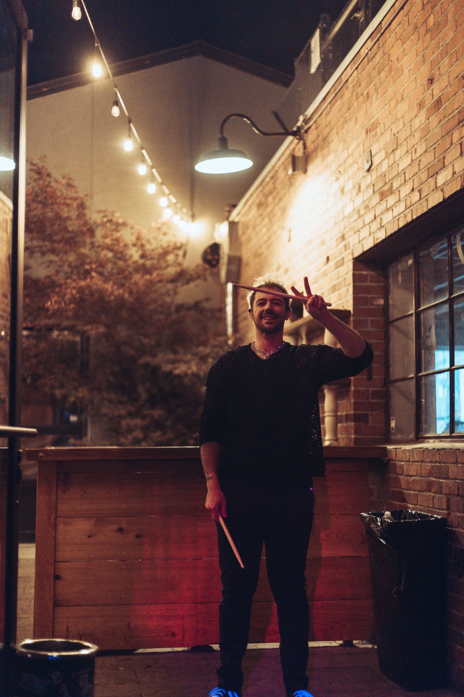 Person in dark clothing stands in a dimly lit courtyard with string lights and brick walls at night.