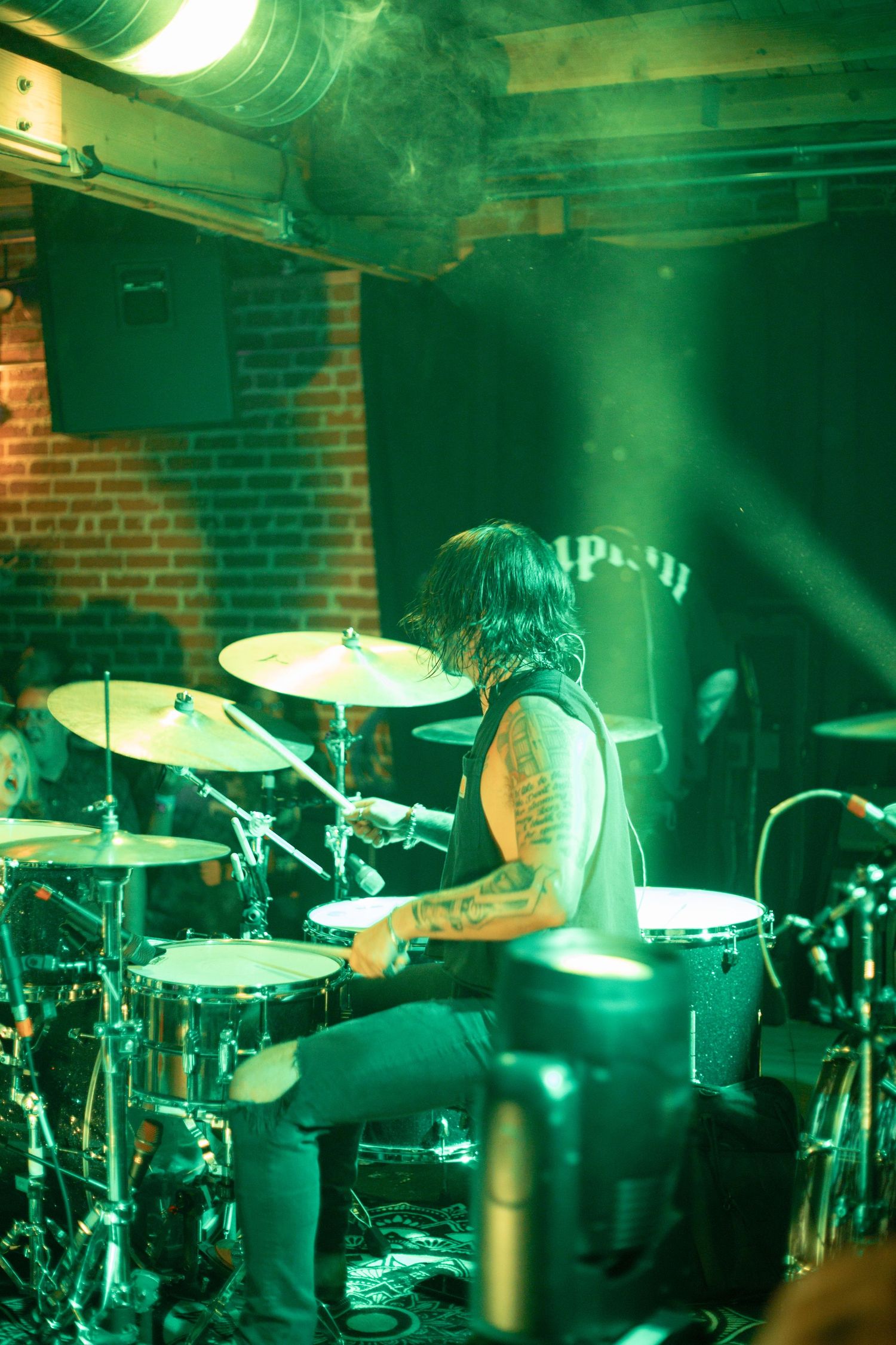 Drummer performing on stage with drum kit under green lighting in brick-walled venue.