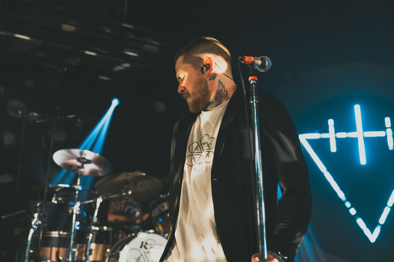 Tattooed male vocalist performing on stage with microphone stand, drums and blue neon lights in background.