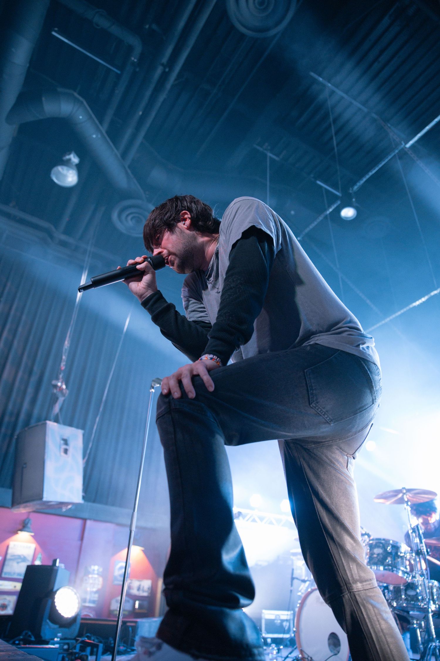Singer performs energetically on stage in a dramatic pose during concert with blue lighting and drum kit visible in background.