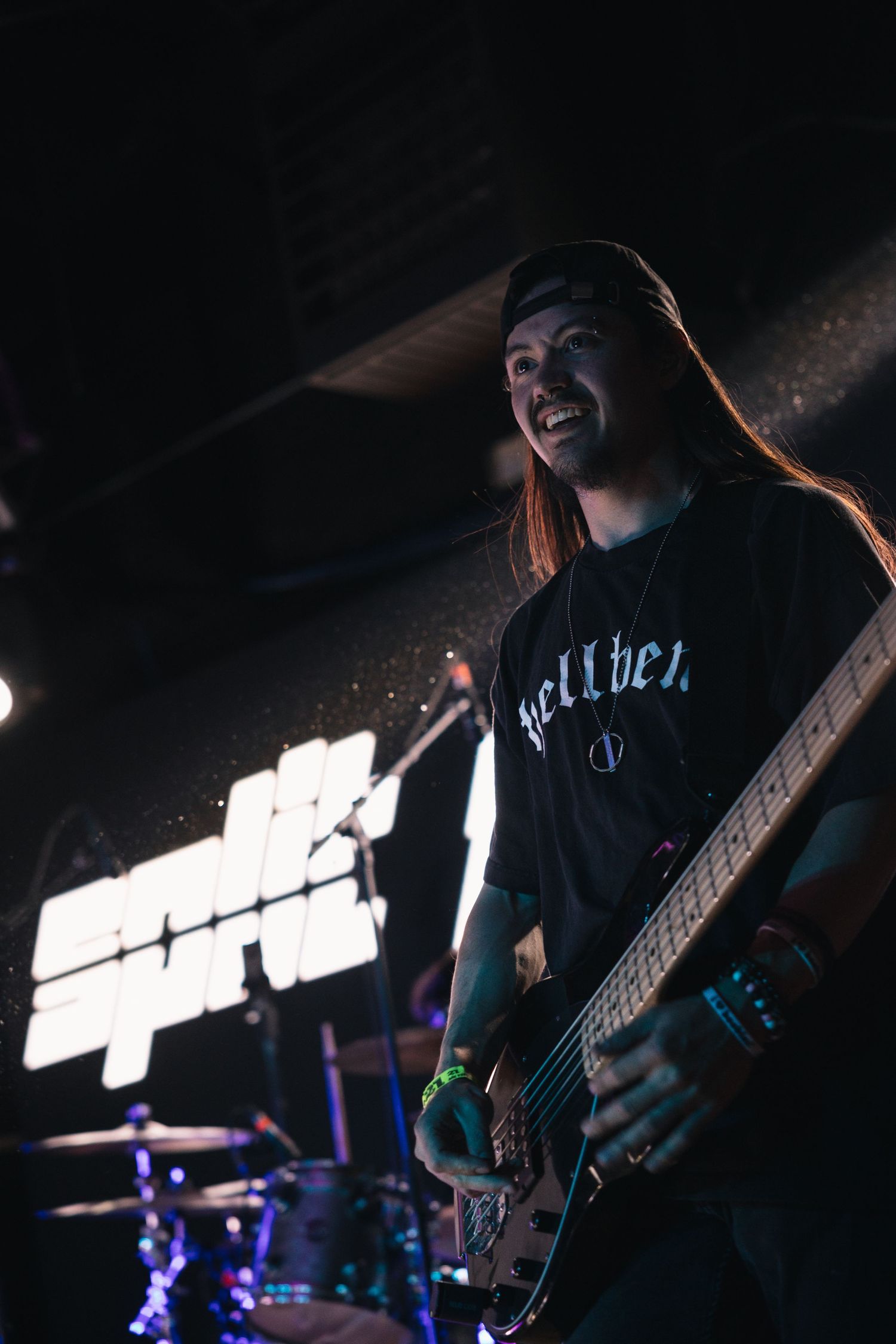 Long-haired guitarist performs on dark stage wearing black t-shirt, smiling while playing bass guitar at live rock concert.
